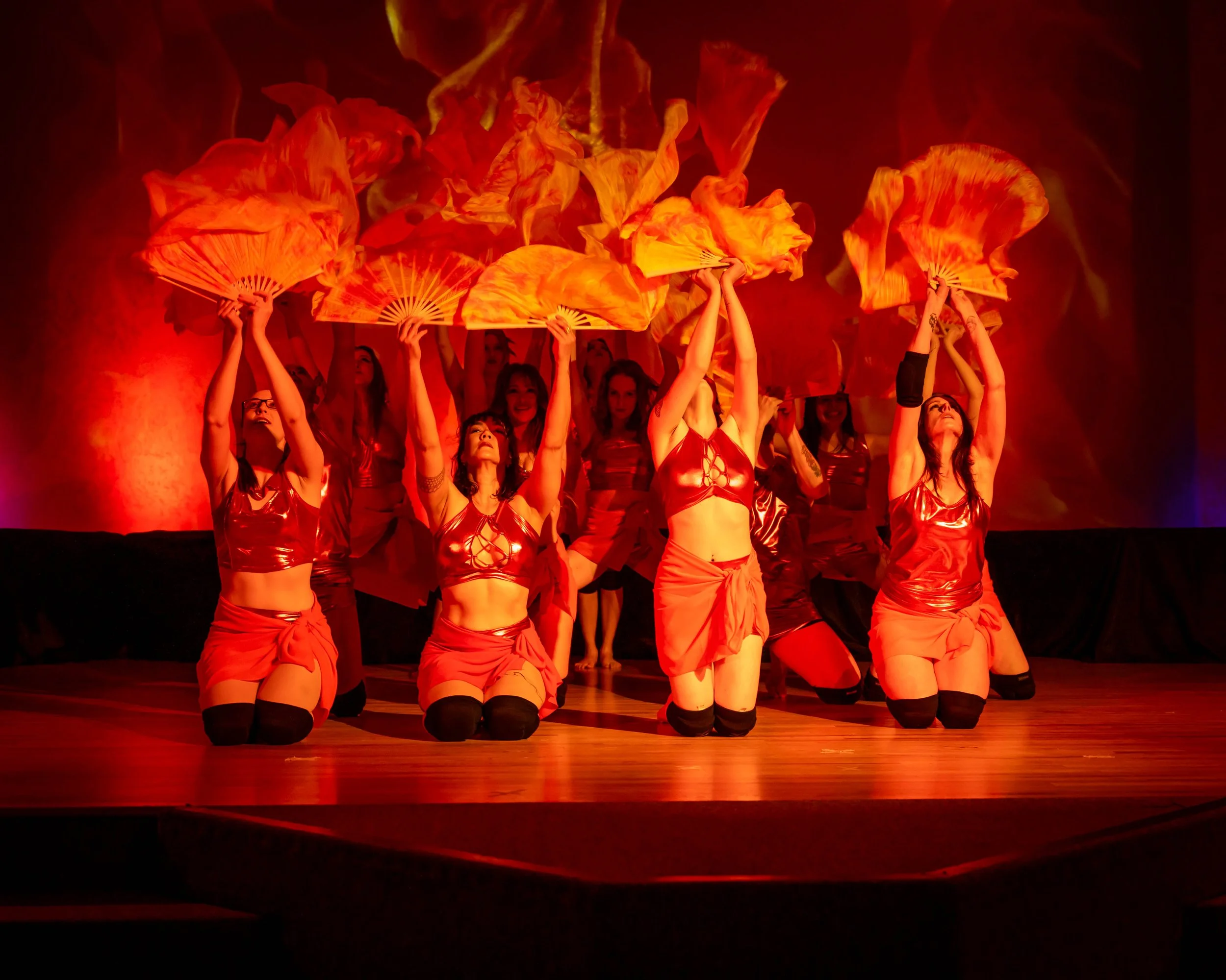 Group of women in red costumes performing a dance with large orange fans on a wooden stage, with red lighting.