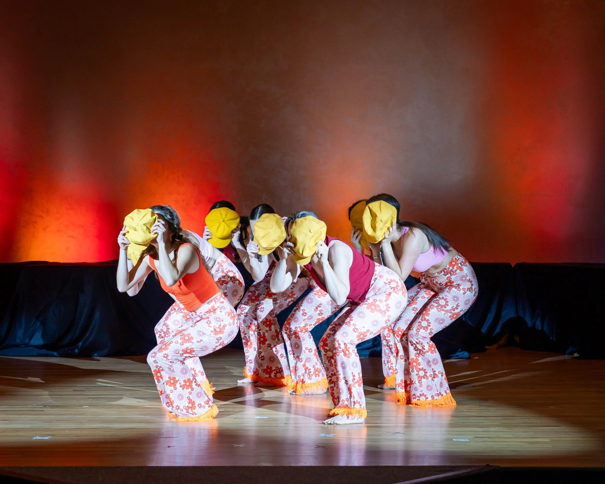 Group of women in floral pants and pink or red tops performing on stage while holding yellow cloths over their faces.