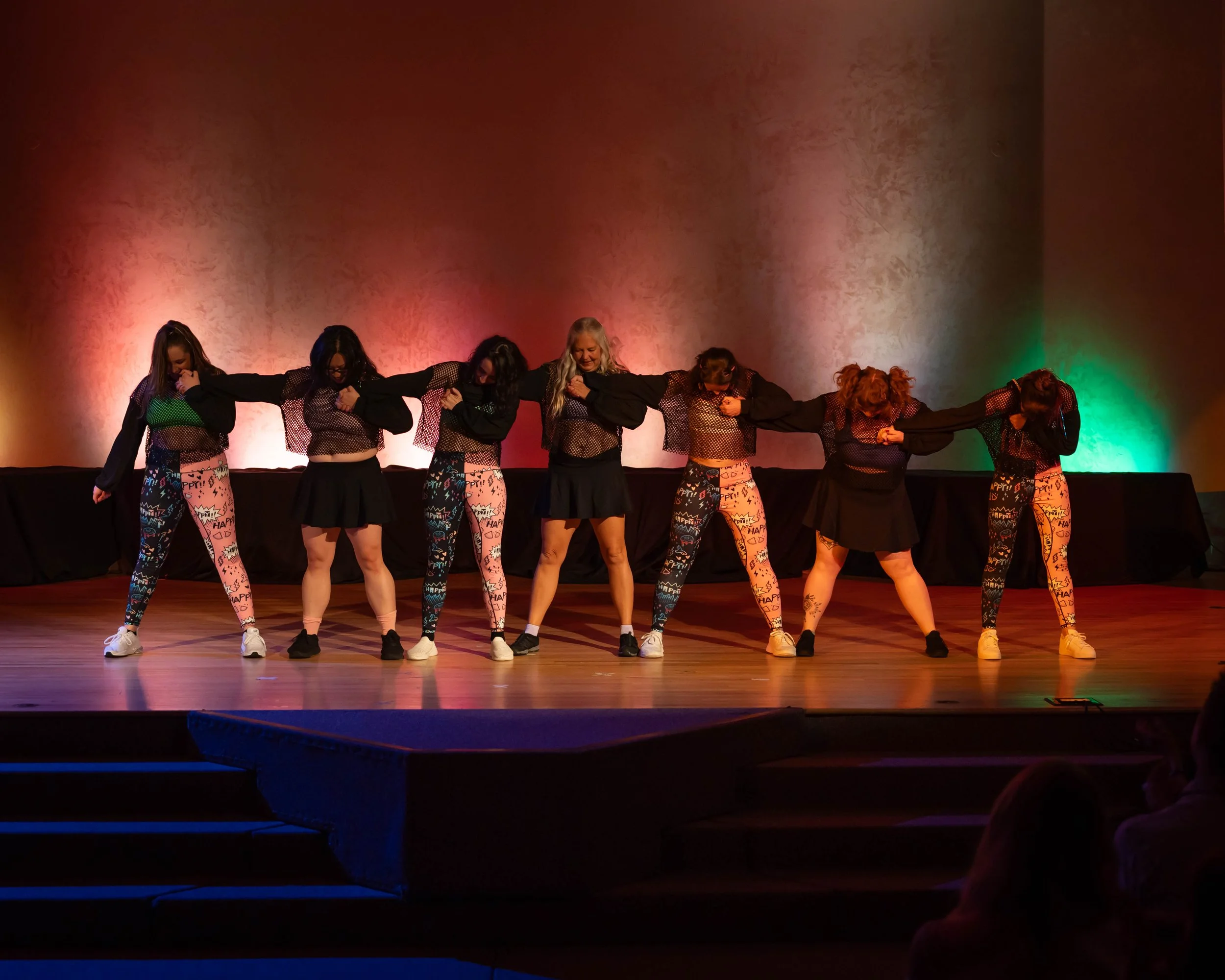 Group of seven women performing a dance on stage, holding hands and leaning to the side, with colorful lighting in the background.