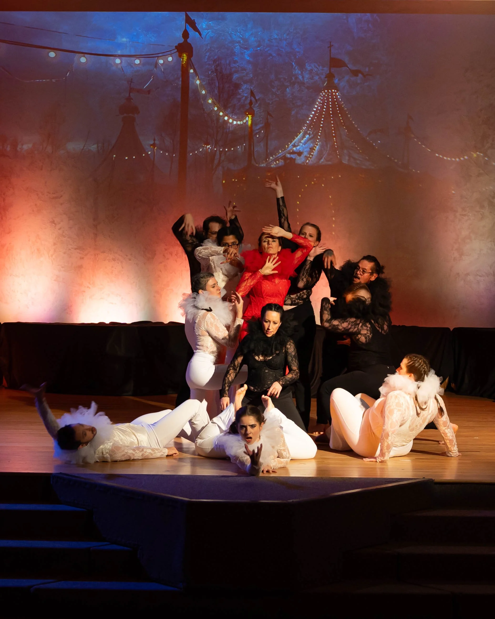 A group of performers in elaborate costumes on a stage, with a circus-themed backdrop featuring tents and flags.