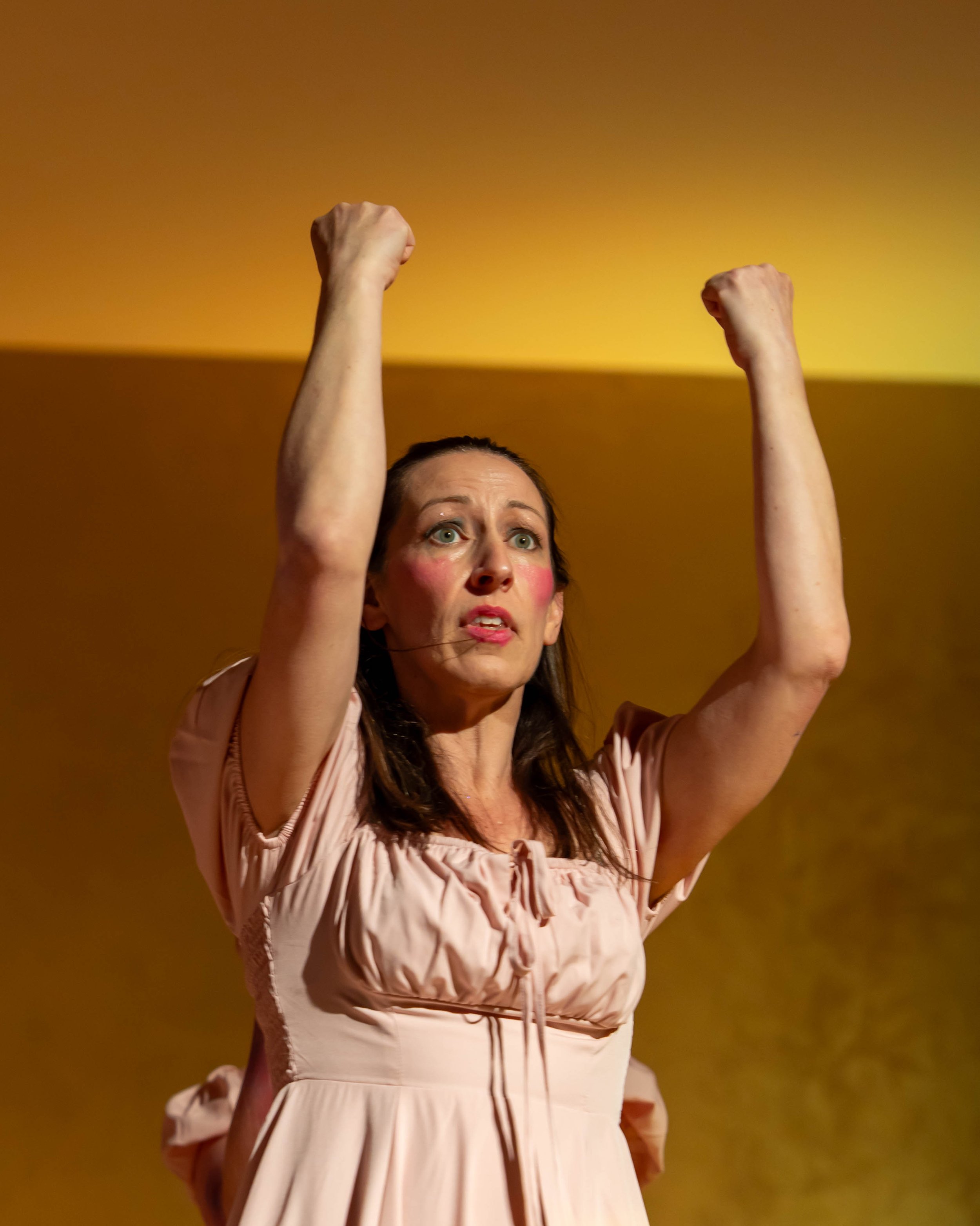A woman with long dark hair and a pink dress, raising both fists in the air, looking determined, against a yellow background.