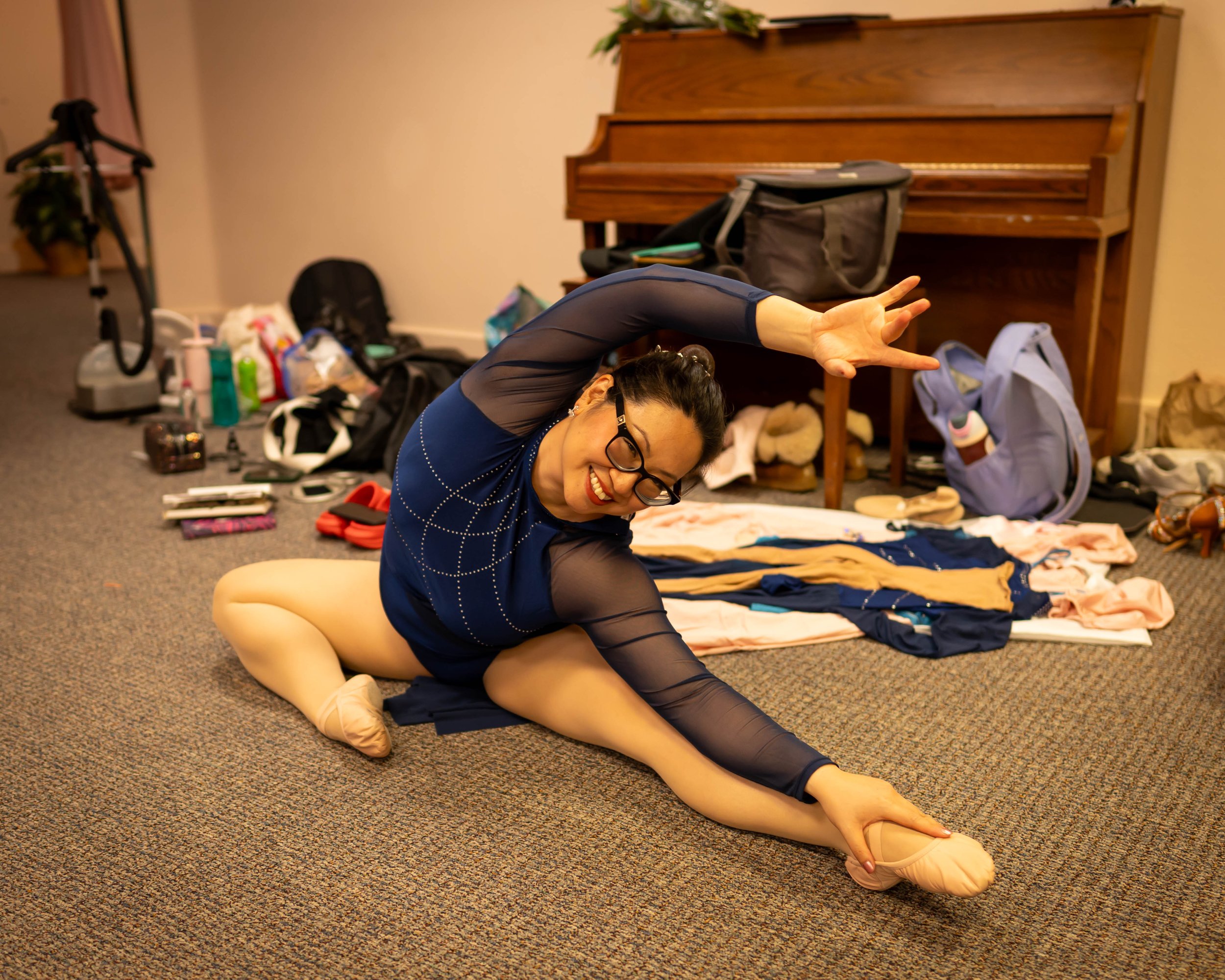A woman in a blue costume stretching on the floor of a room with various clothes and bags around her, and a piano behind her.