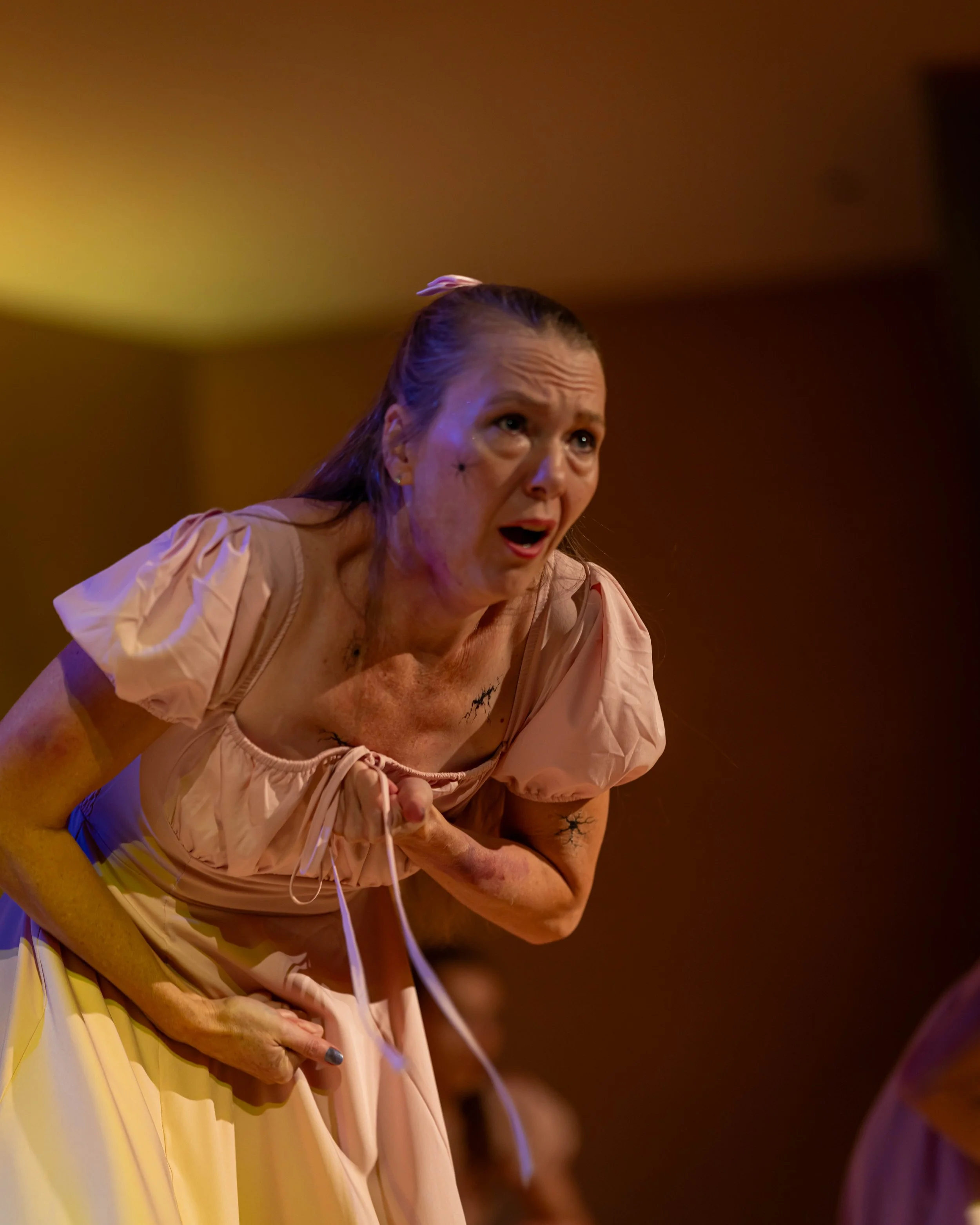 A woman with long hair, wearing a pink dress, appears distressed and is clutching her stomach as she bends forward in a moment of emotion.