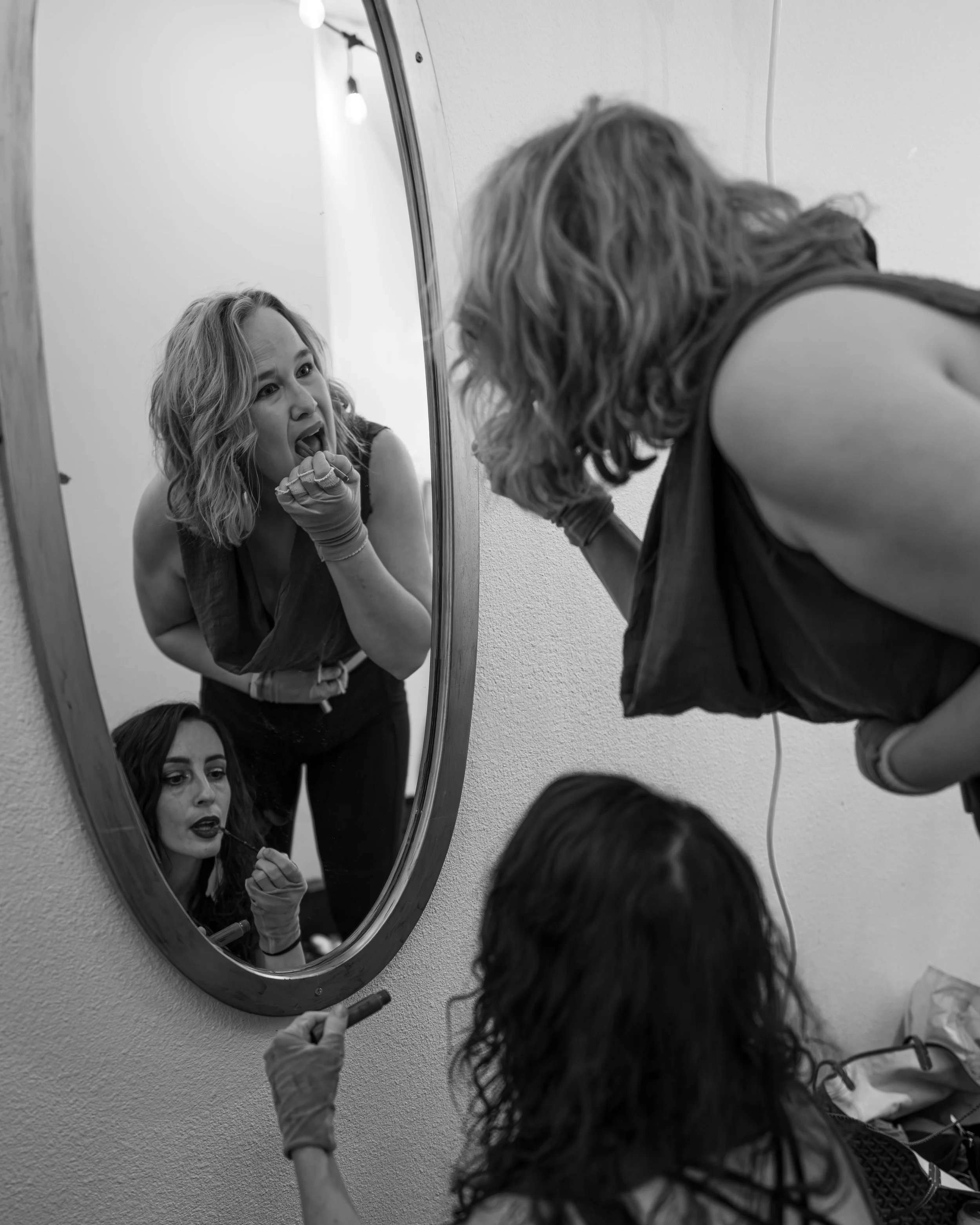 Two women, one applying lipstick and the other looking into a mirror, as seen from outside the mirror, in black and white.