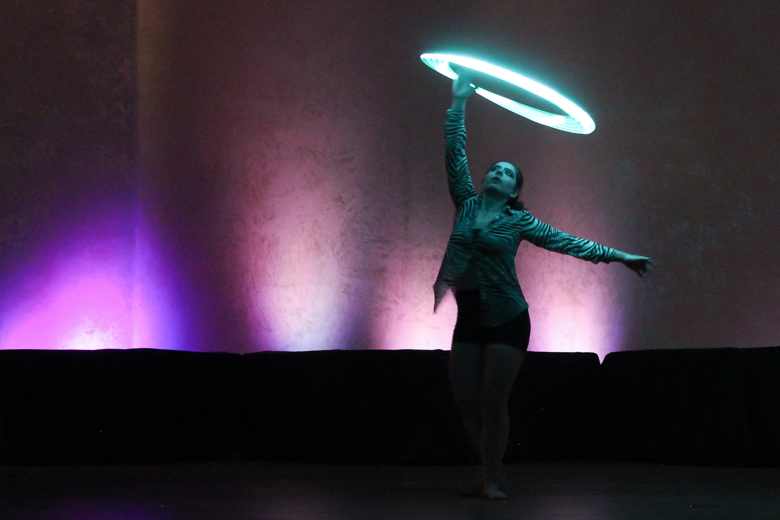 A woman performing with a glowing hula hoop in a dark room with colorful lights on the wall.