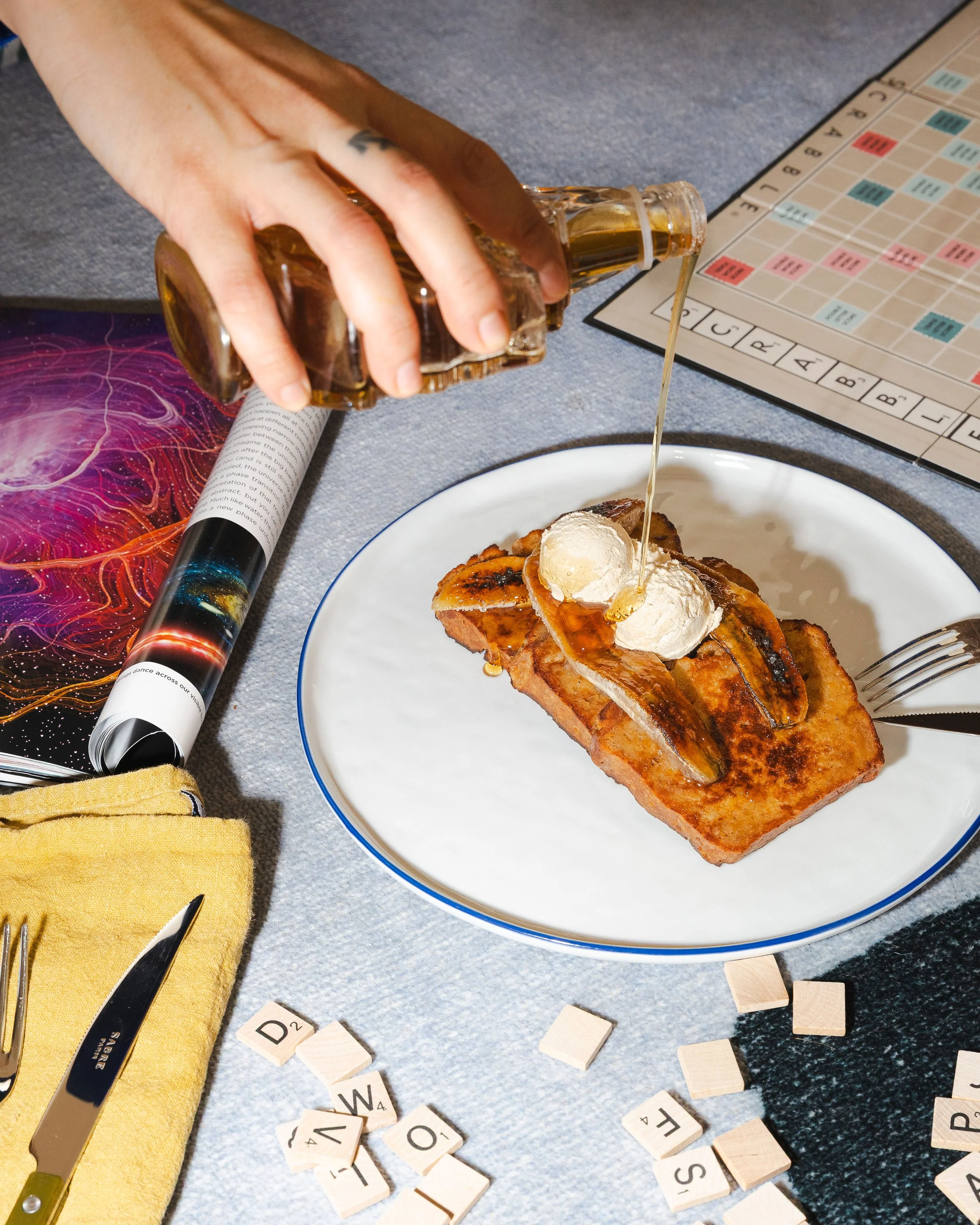 A person pouring syrup on French toast topped with espresso honey butter on a white plate, with Scrabble tiles, a fork, a knife, a board game, a magazine, and a yellow napkin on a blue table.