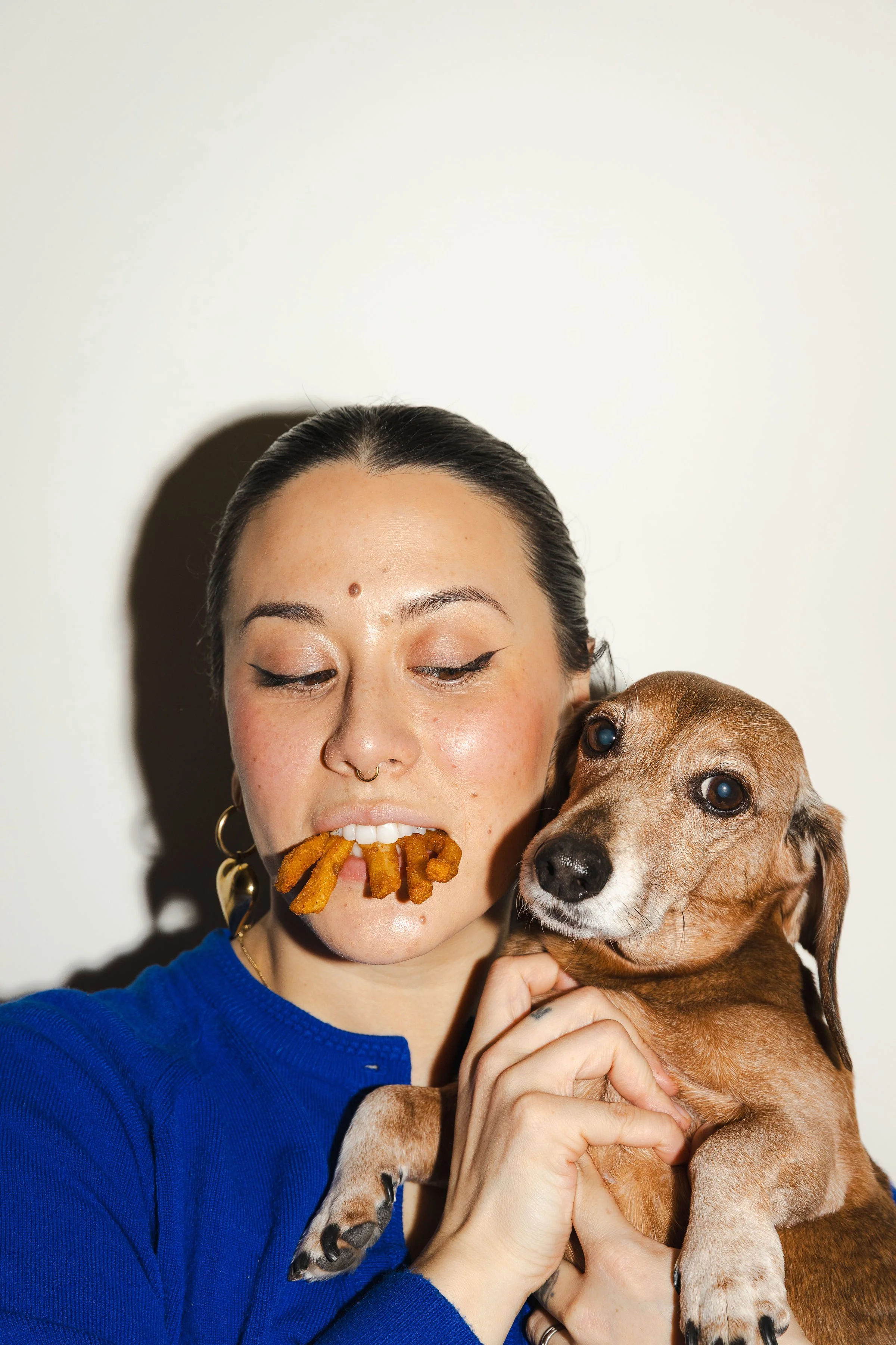 A woman holding a dachshund dog, with some fries sticking out of her mouth.