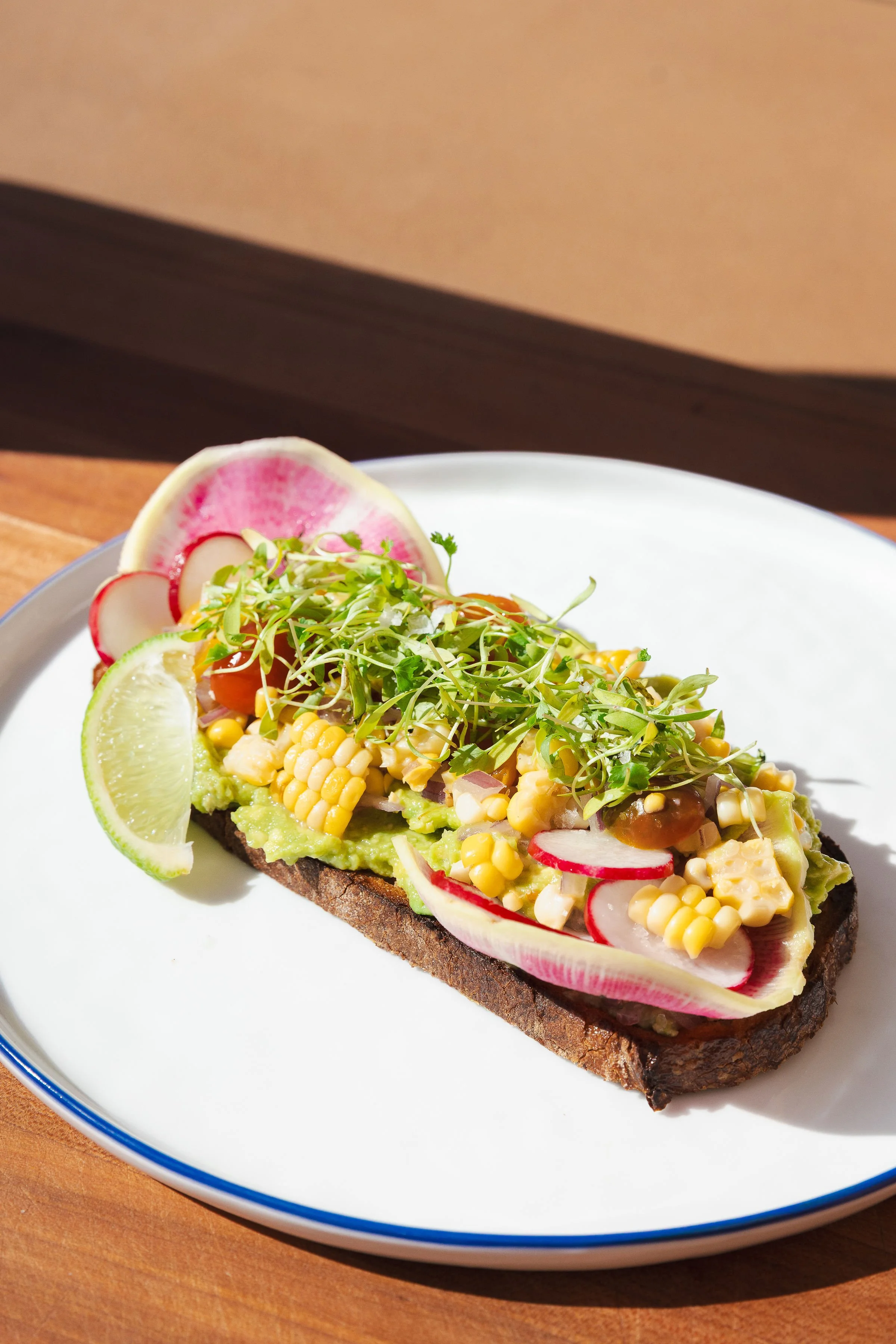 Open-faced avocado toast topped with microgreens, sliced radishes, corn, and a lime wedge on a white plate.