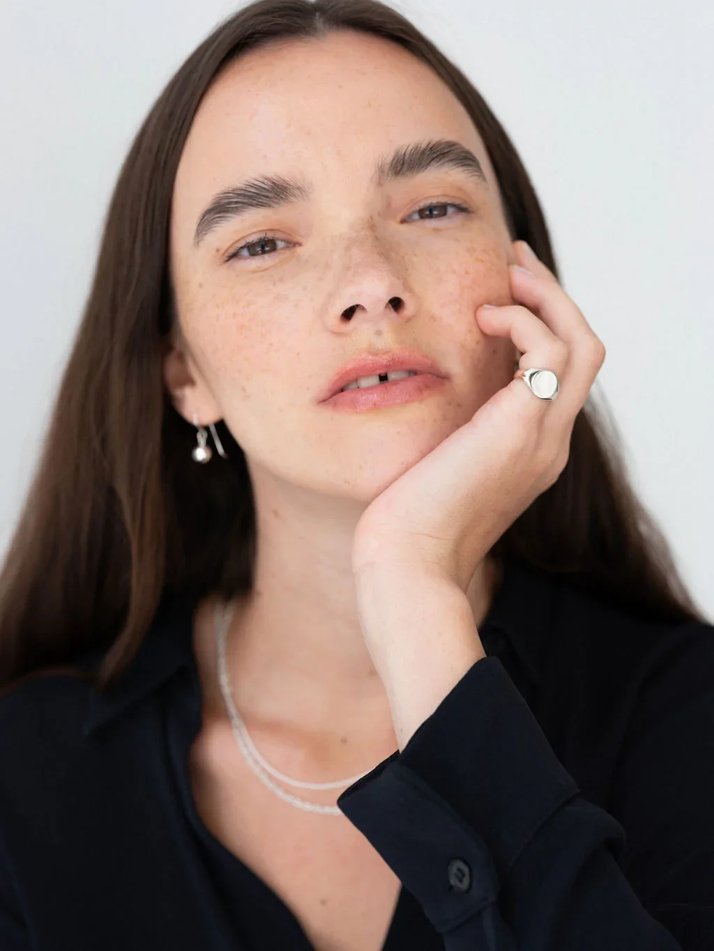 A young woman with long brown hair, freckles, and light eyes, resting her head on her hand, wearing earrings, a layered necklace, and a black shirt, with a neutral background.