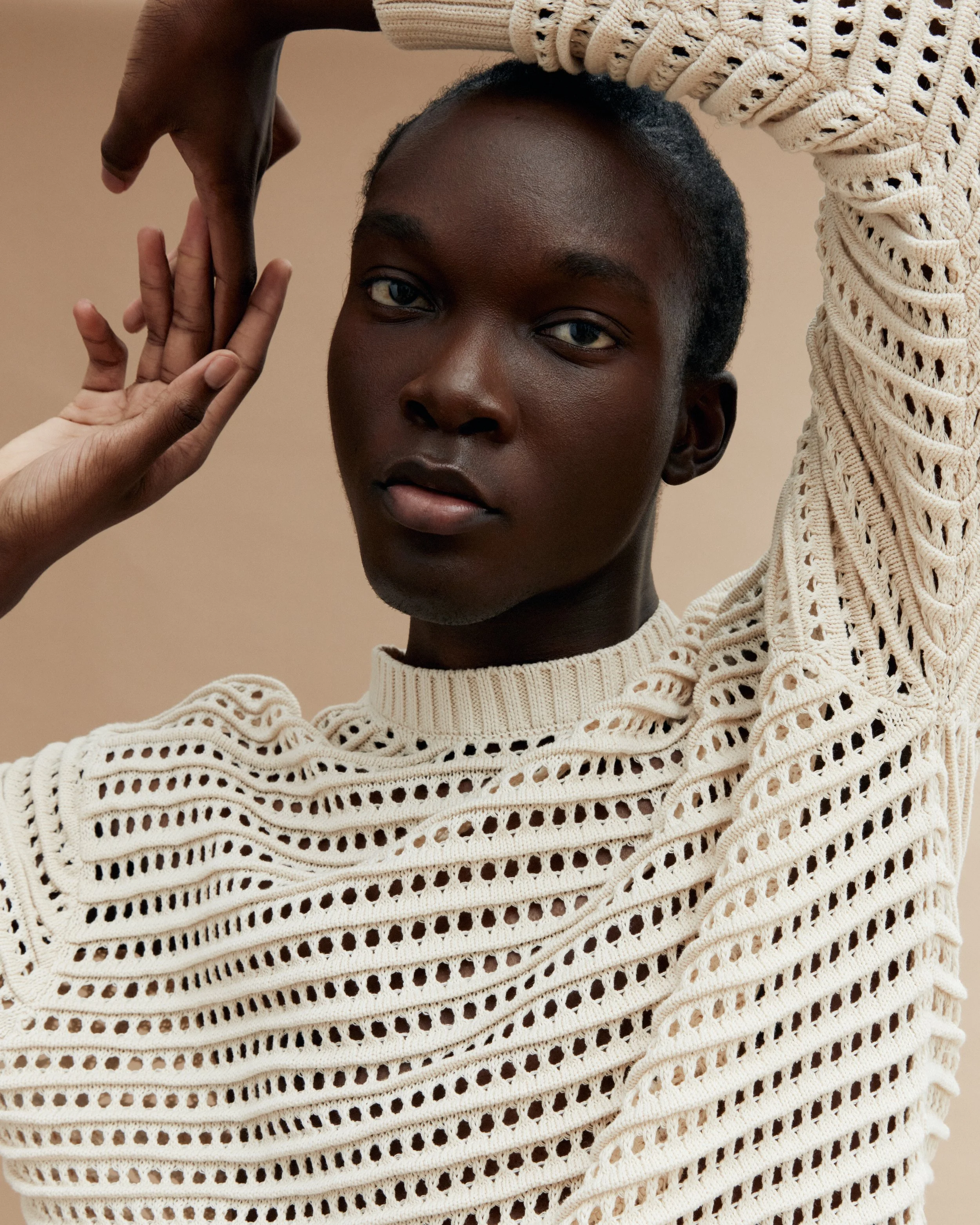 A young black woman with short hair wearing a cream-colored, open-knit sweater, holding her arm above her head and gently touching her face with her left hand, against a plain beige background.