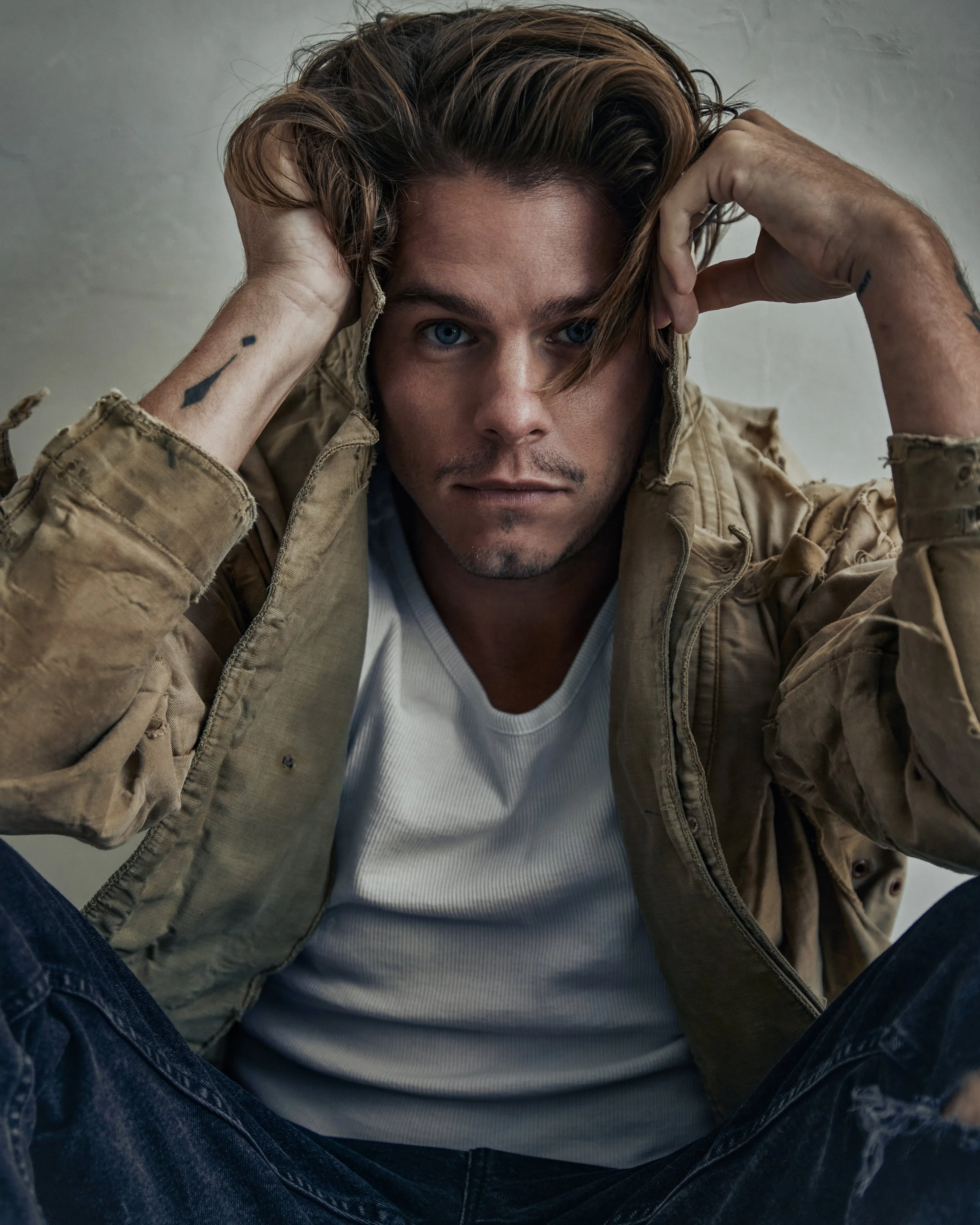 A man with blue eyes, brown hair, and a light beard, sitting with his legs crossed, wearing a white shirt and a beige jacket with distressed jeans, looking into the camera in a close-up shot.