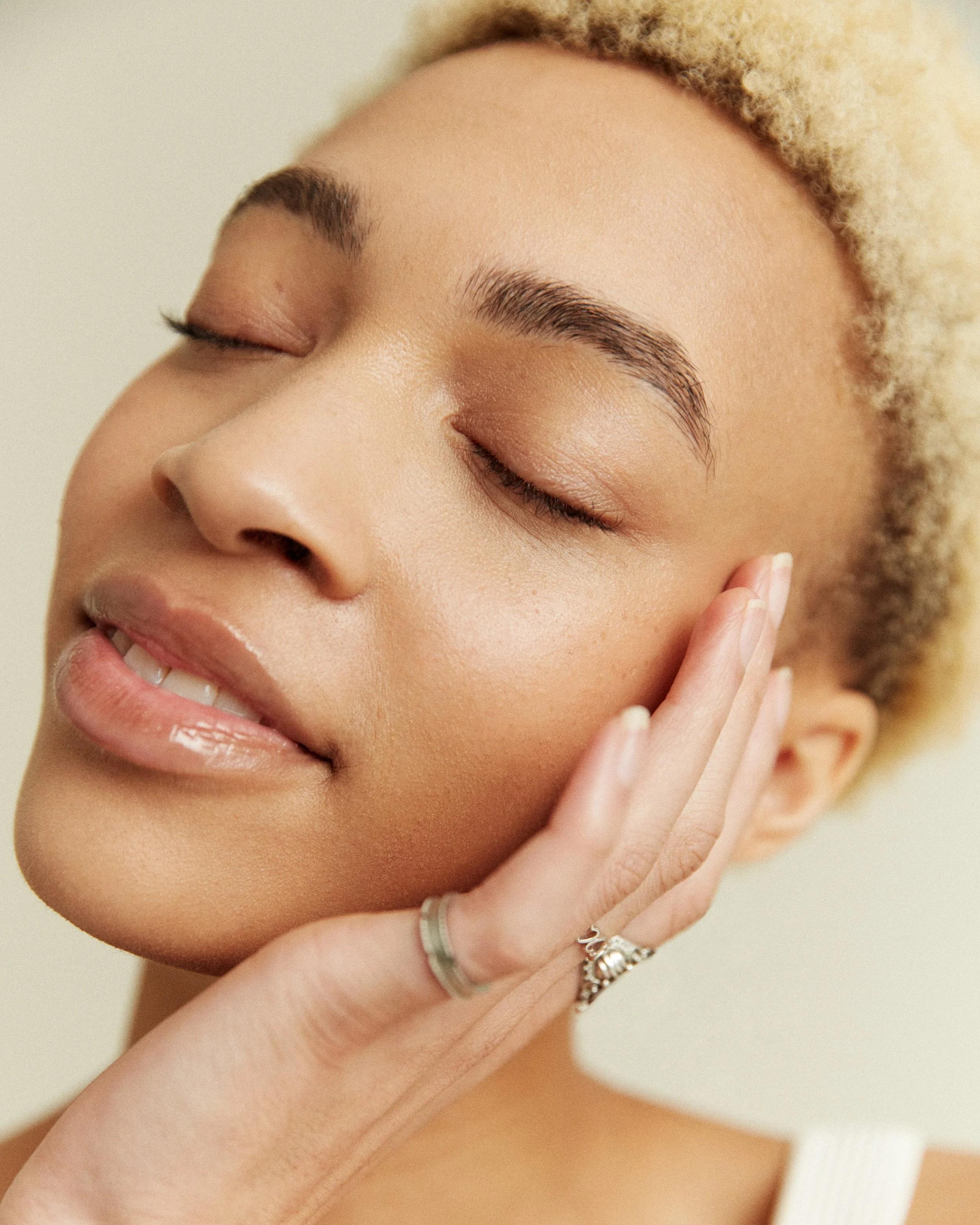 Close-up photo of a woman with blonde curly hair and closed eyes, showing her face, hand with rings on fingers, and smooth complexion.
