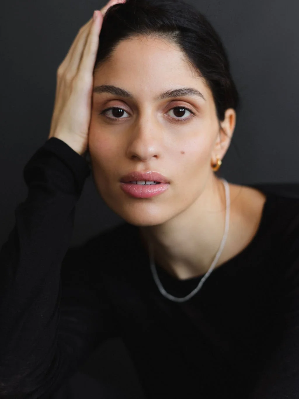 A young woman with dark hair and light skin, staring directly at the camera, holding her head with her right hand, against a dark background, wearing a black top and gold jewelry.