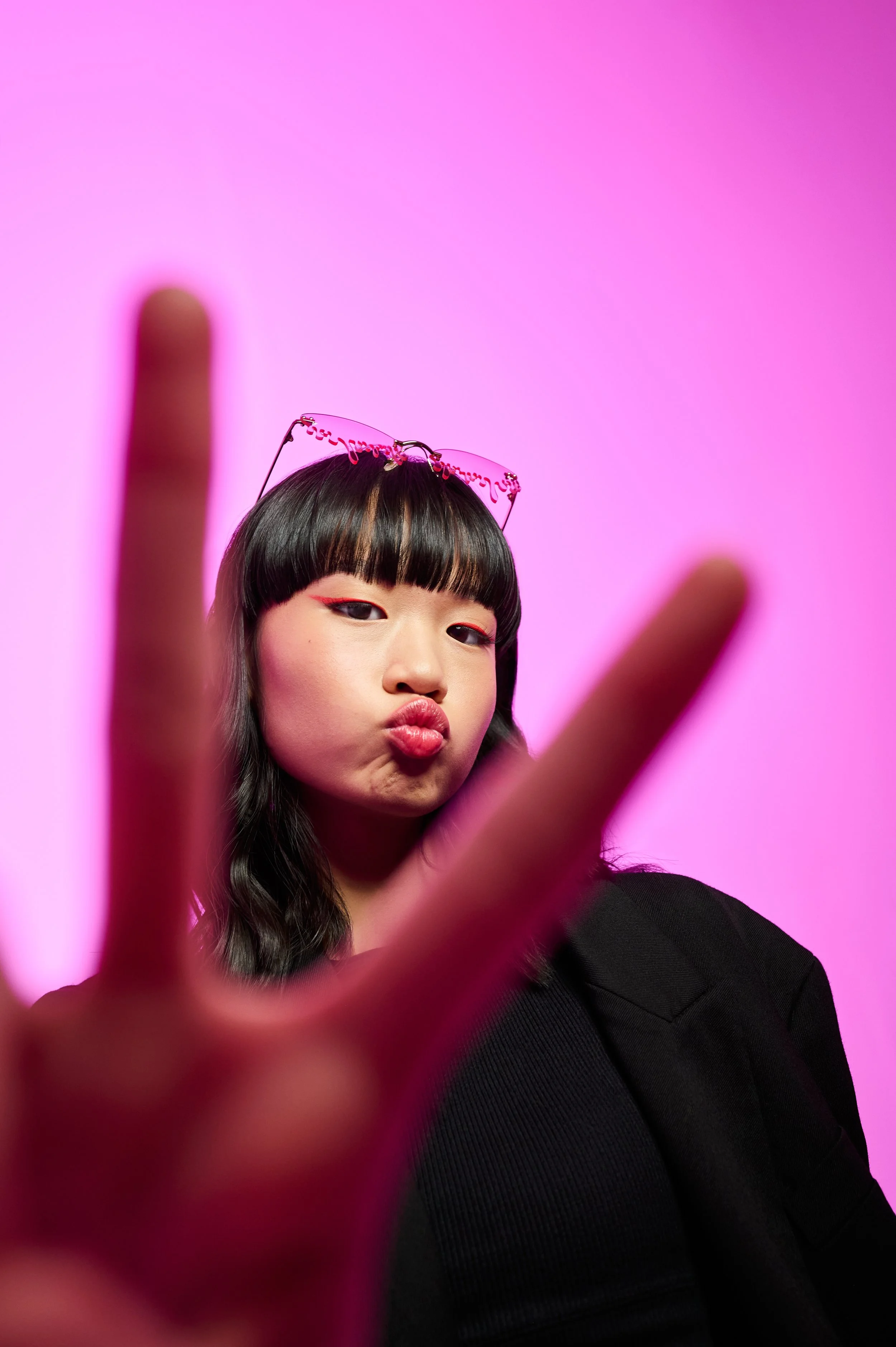 A young woman with dark hair and bangs wearing pink sunglasses on her head, making a duck face, with a blurred finger in the foreground and a pink background.