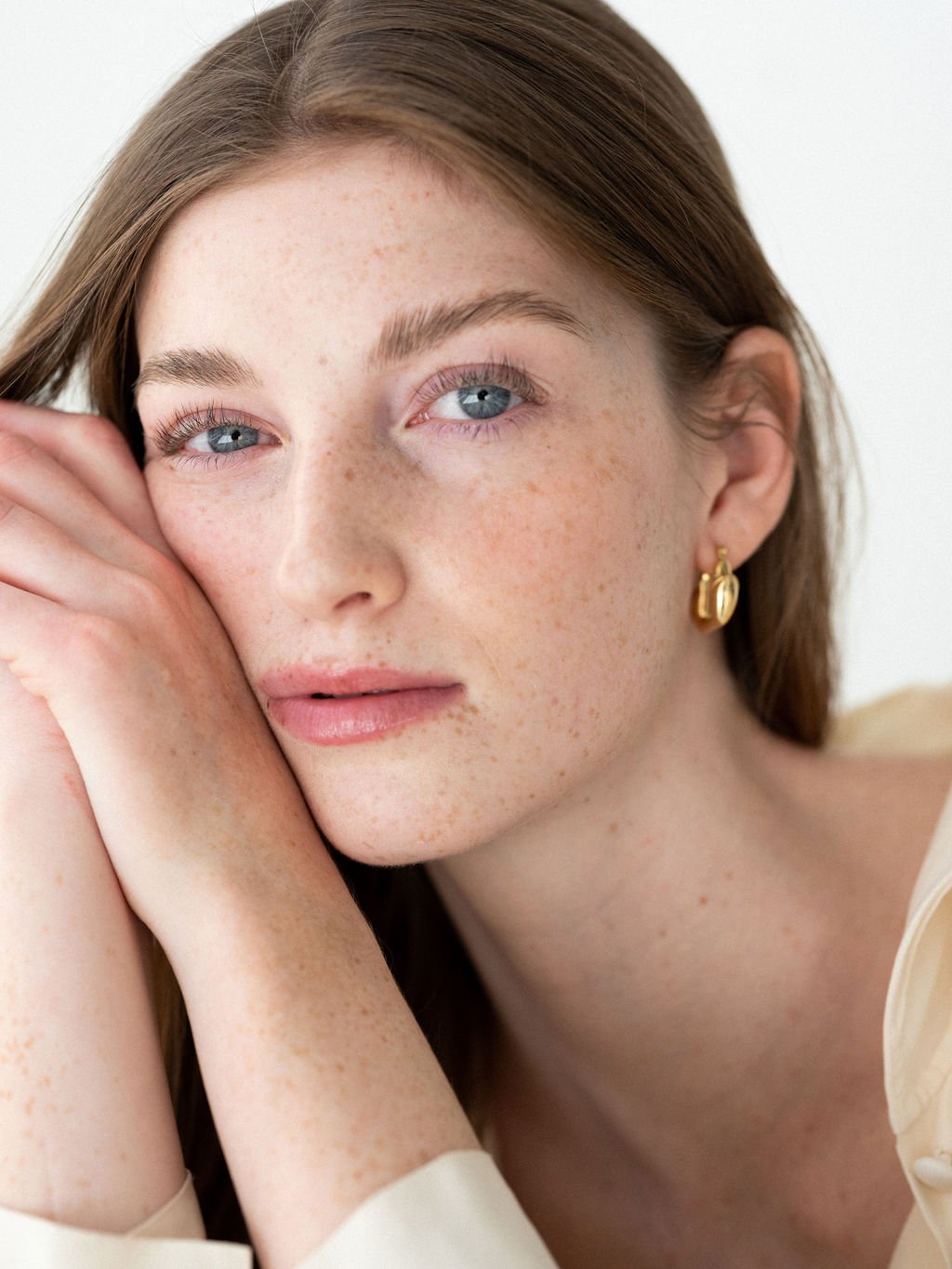 close-up of a woman with red hair and blue eyes, resting her face on her arms, wearing gold earrings, with clear skin and freckles, looking softly at the camera.