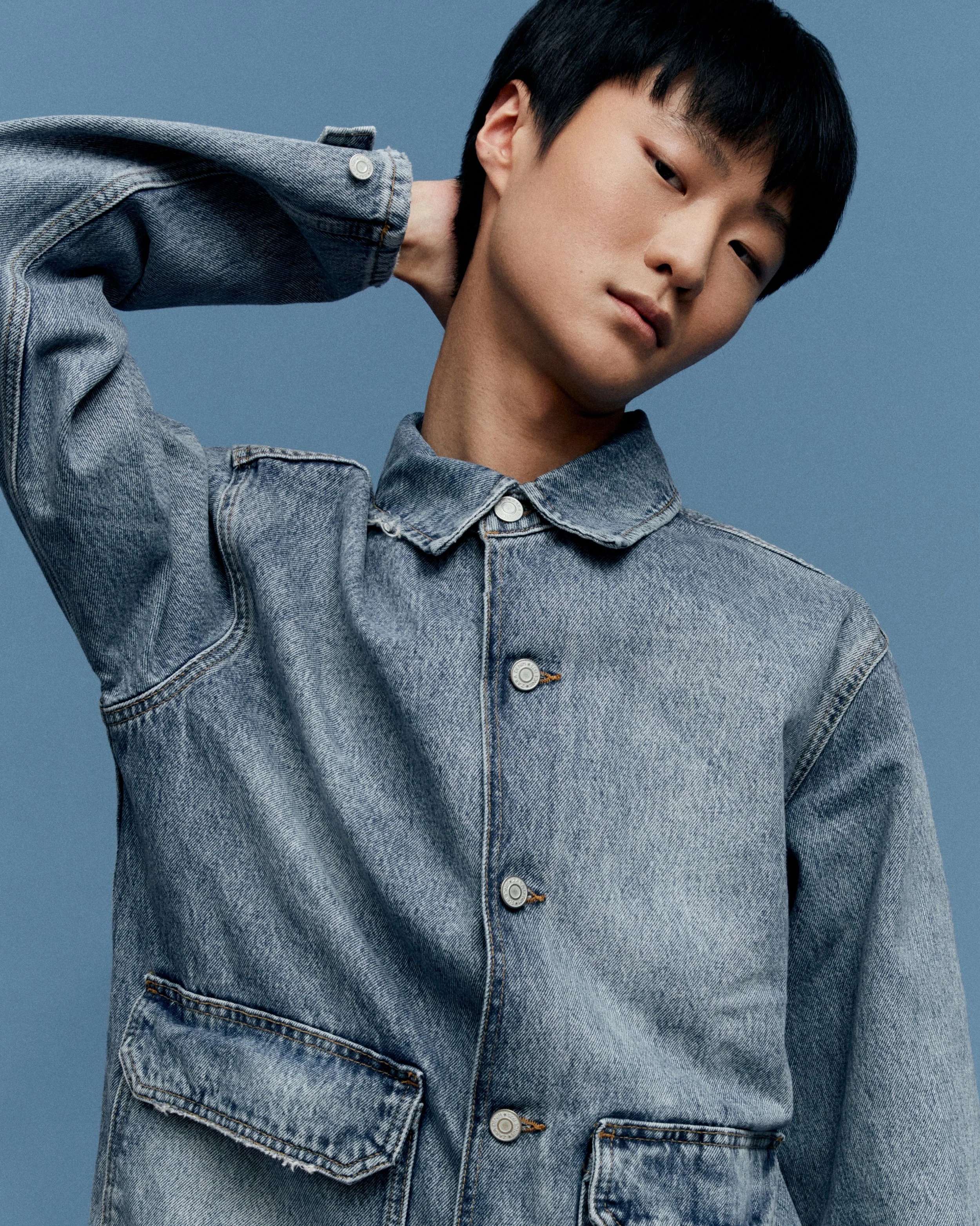 Close-up of a person with short black hair wearing a blue denim jacket against a solid blue background.