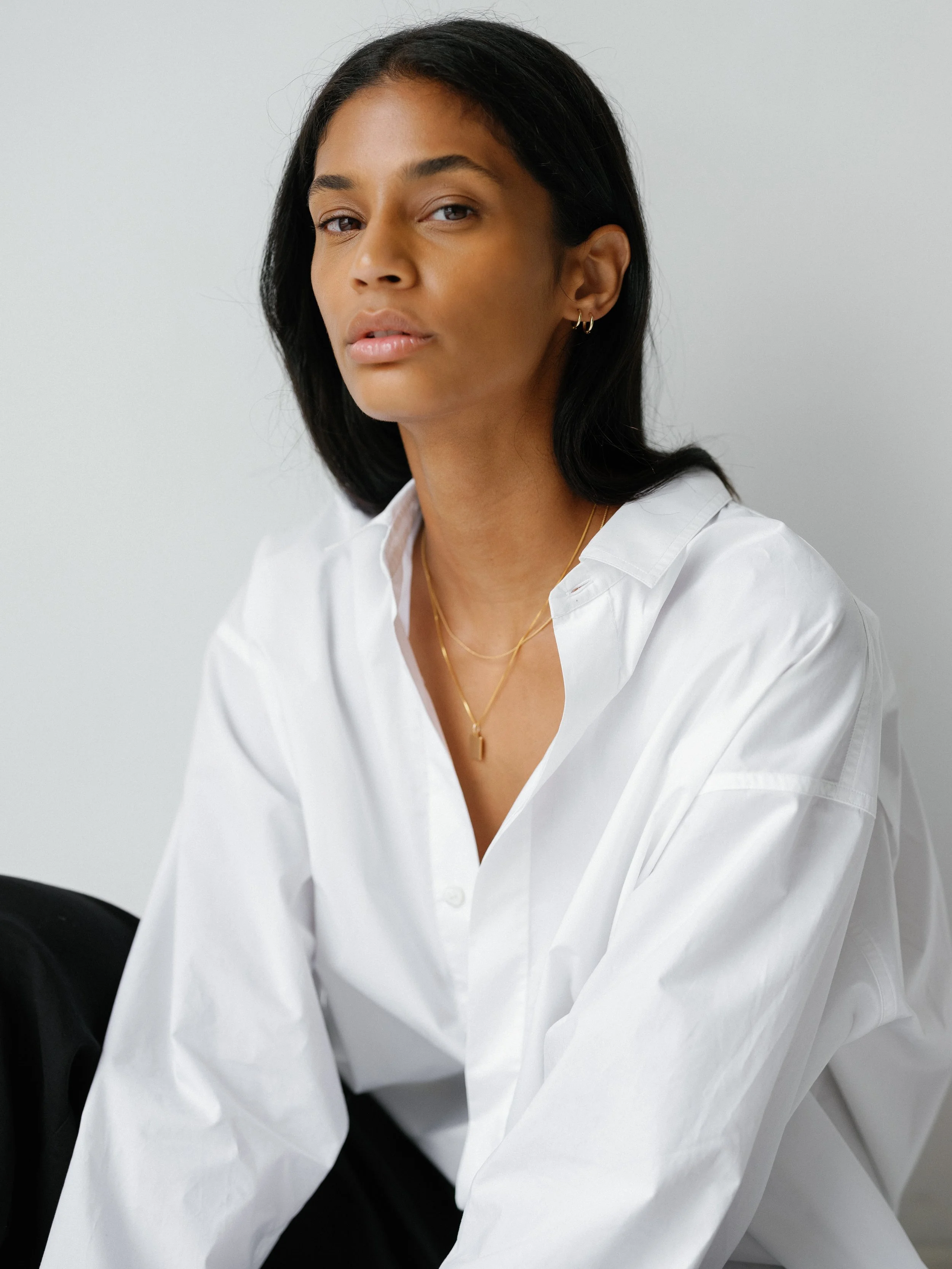 A woman with black hair wearing a white shirt, gold jewelry, and earrings, sitting against a plain light gray background.