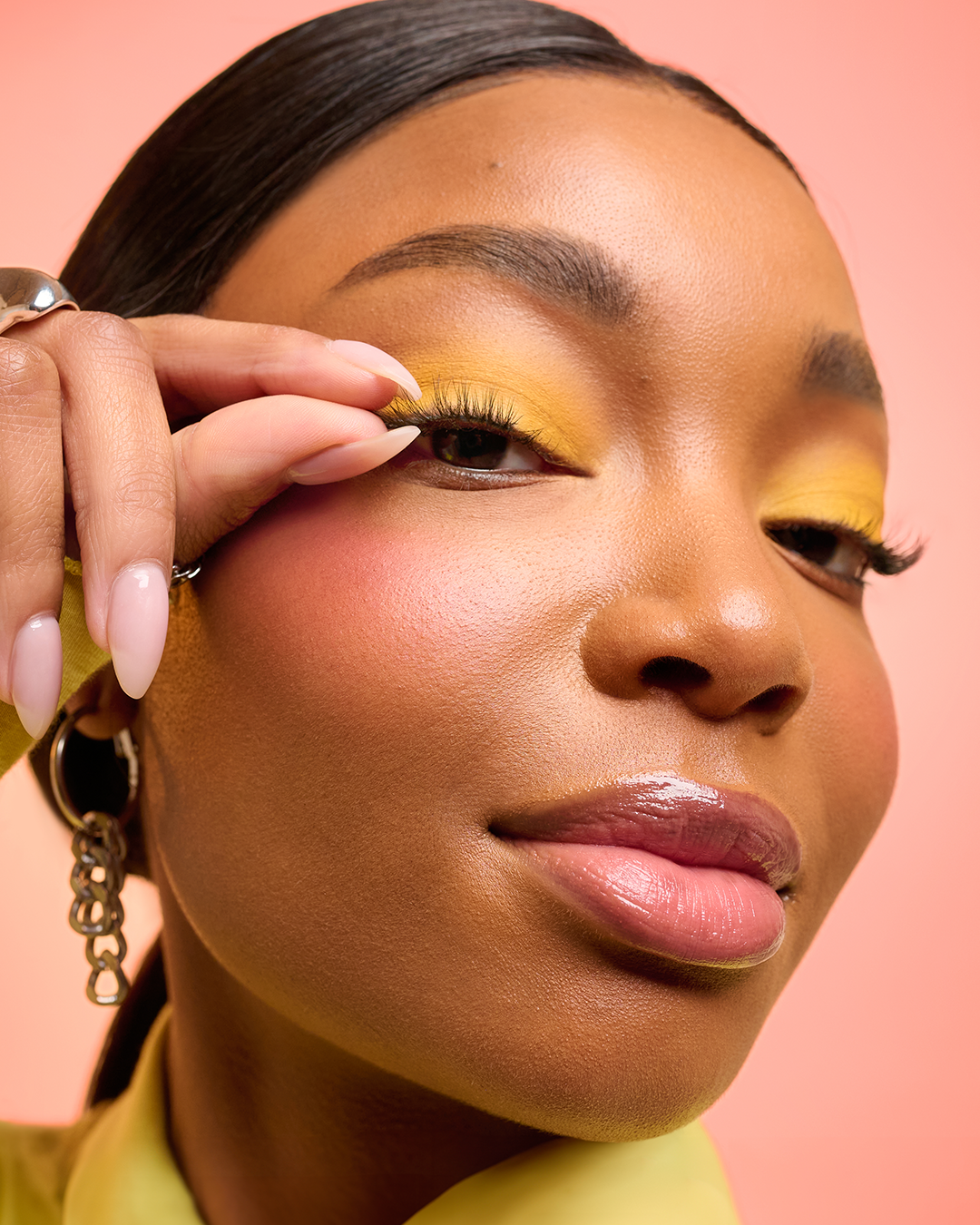 Close-up of a woman with yellow eye makeup, glossy lips, and earrings, touching her eye with her left hand against a pink background.
