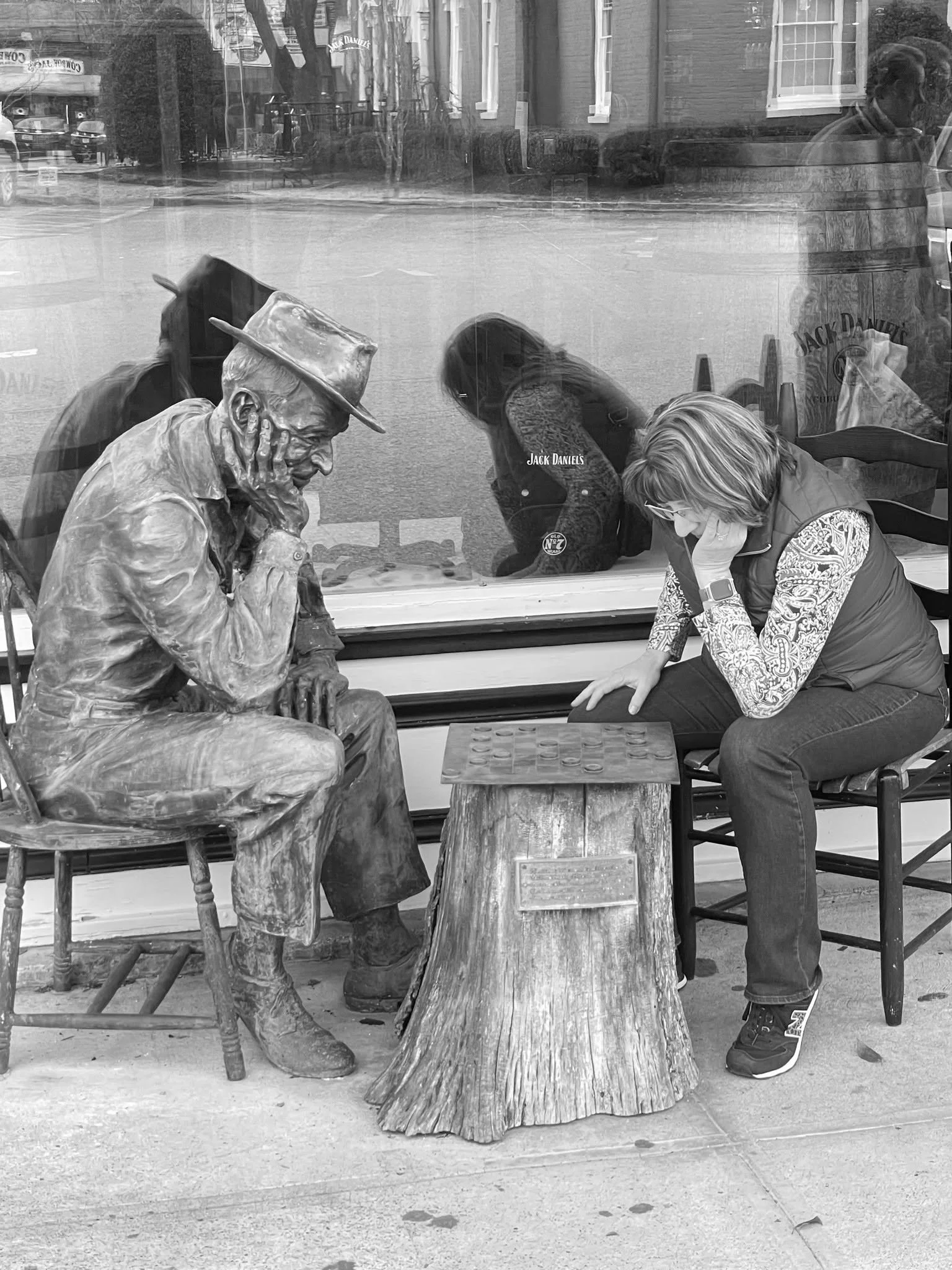 A street scene featuring a bronze statue of Jack Daniel’s sitting on a chair with his chin resting on his hand. Next to the statue, Katrinka, is engaged in a game of checkers on a small wooden table.