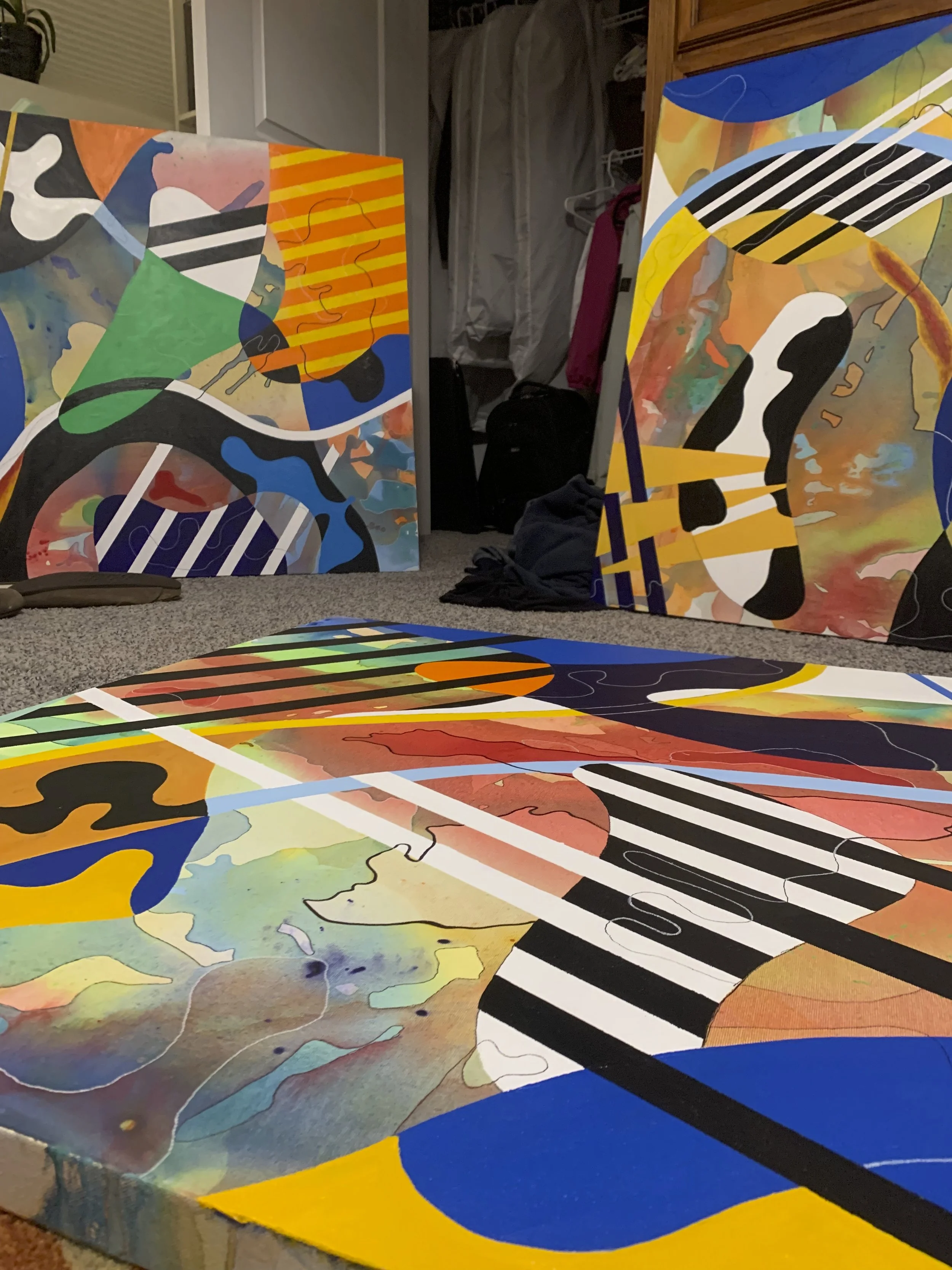 Abstract colorful paintings leaning against a wall in a room, with other artworks and objects visible in the background.