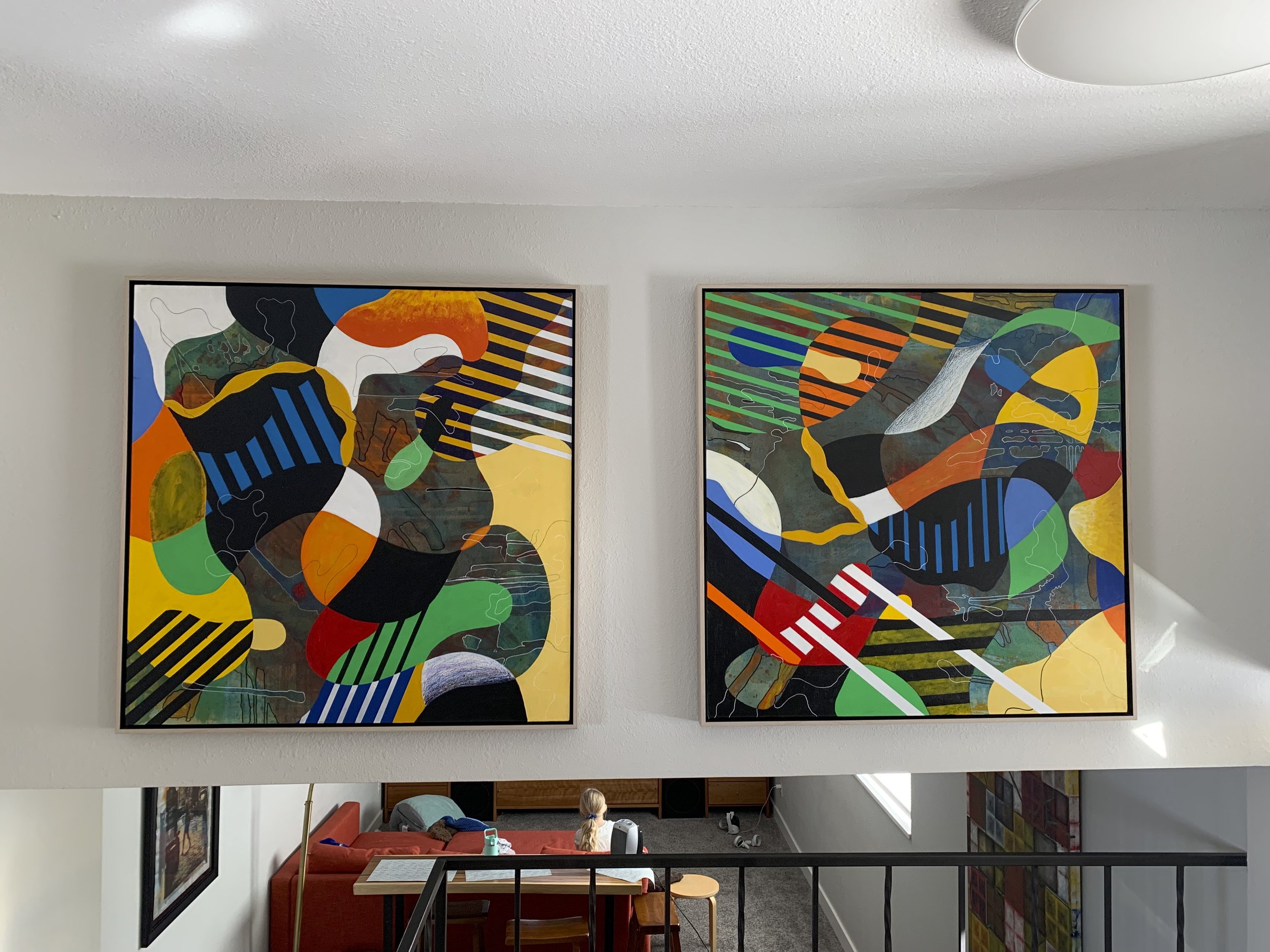 Two colorful abstract paintings hang on a white gallery wall. A sitting area with a red couch and children sitting on the floor is visible below the paintings.