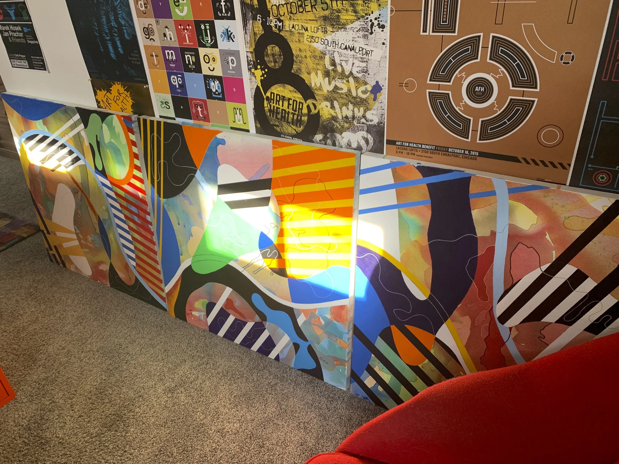 Colorful art display with various posters and designs on panels, including maps, text, and graphic elements, with a red upholstered chair visible in the foreground.