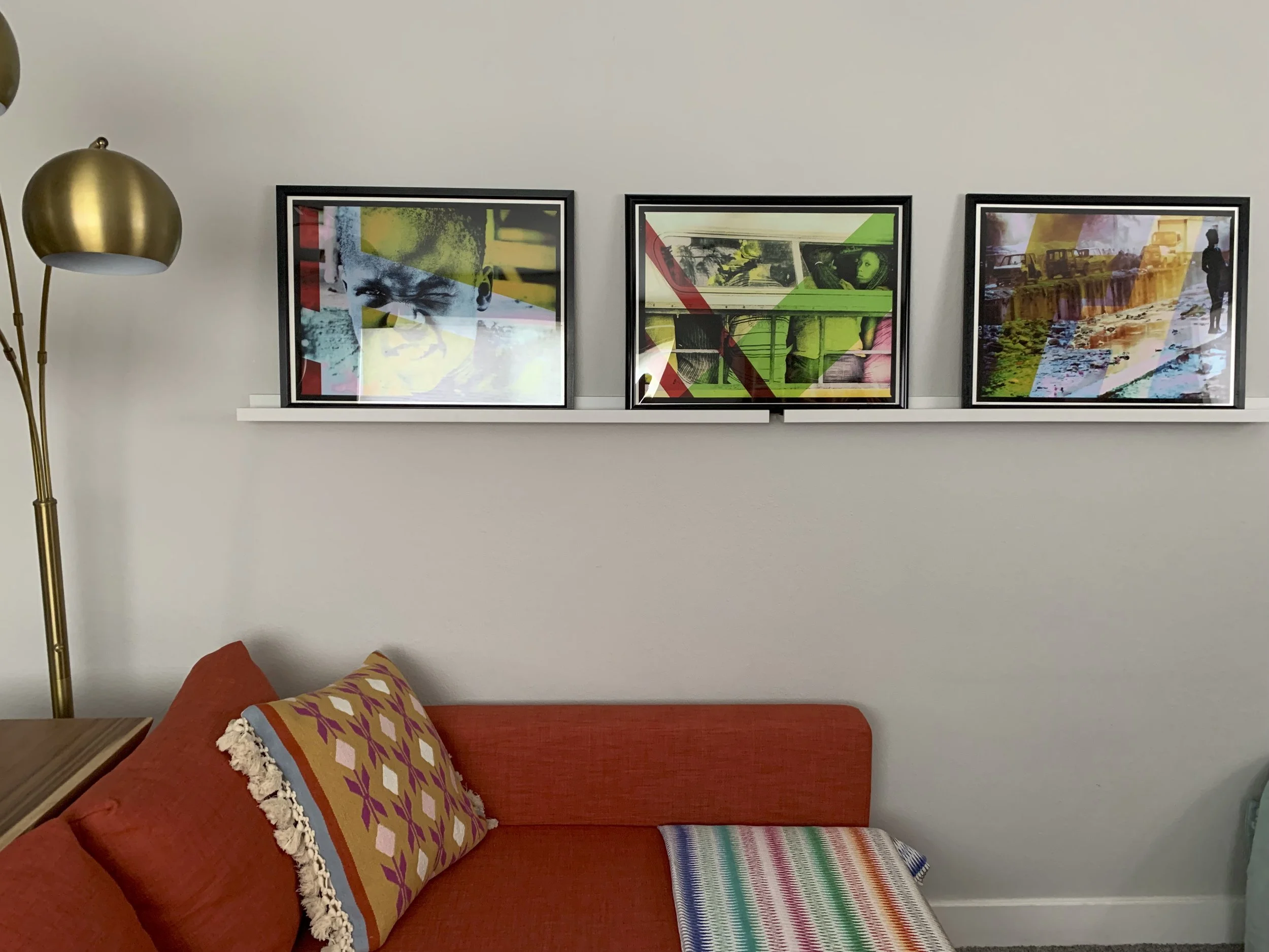 A living room wall decorated with three colorful framed abstract art prints, placed on a white shelf above an orange sofa with patterned and striped throw pillows, with a gold floor lamp on the left.