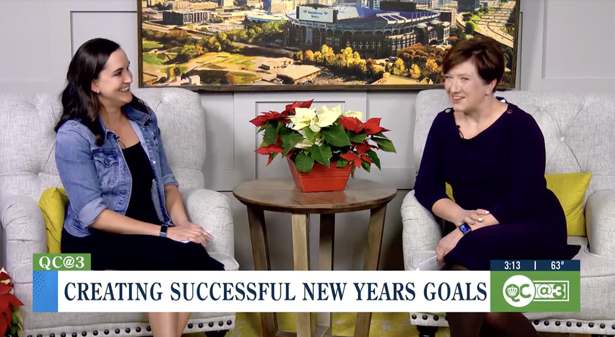 Two women sitting on chairs with a table and potted poinsettia plant between them, discussing New Year's goals on a TV segment. A large picture of a cityscape is in the background.