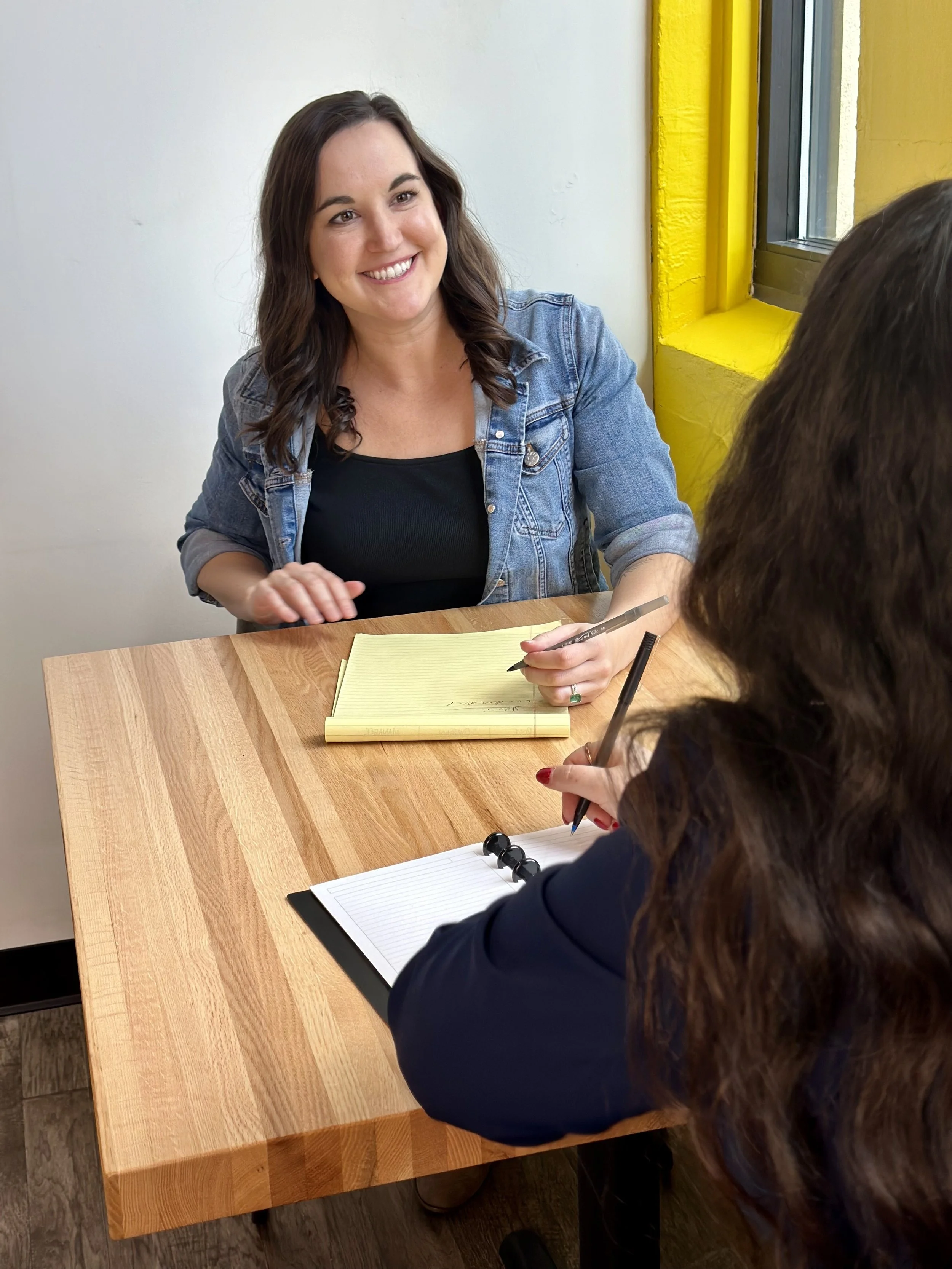 Leadership Coach Brittany Canaski sitting at table smiling at person sitting across from her. They are both writing in notepads and we can just the back of the other person's head with brown curly hair.