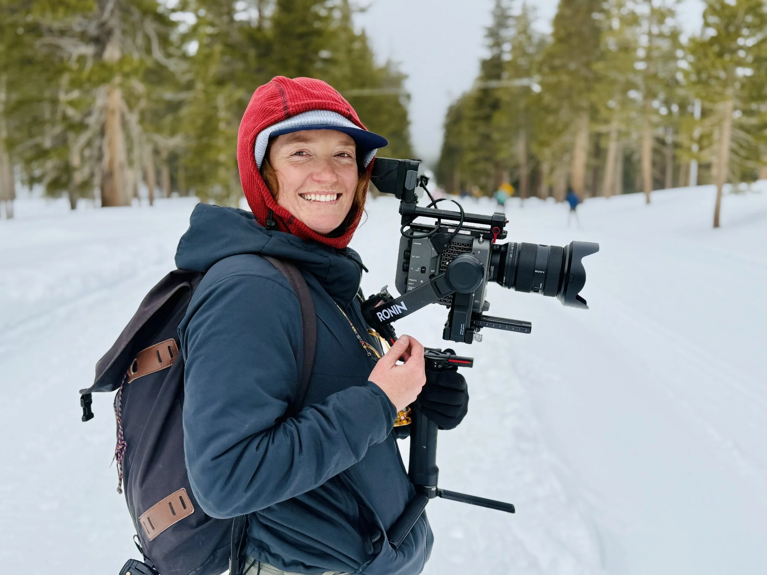 A woman with a red and blue knit hat, brown hair, and a blue jacket, smiling as she holds a professional video camera on a snowy landscape with trees in the background.