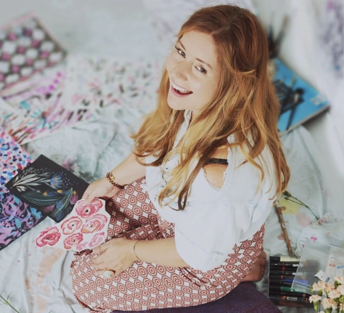 Woman sitting on the floor surrounded by colorful artwork and holding a floral painting. She is smiling, with long hair and wearing patterned pants and a white top.