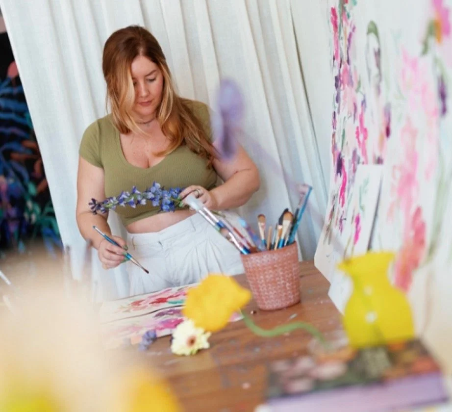 Woman painting floral artwork in a studio with paintbrushes and flowers on a table.