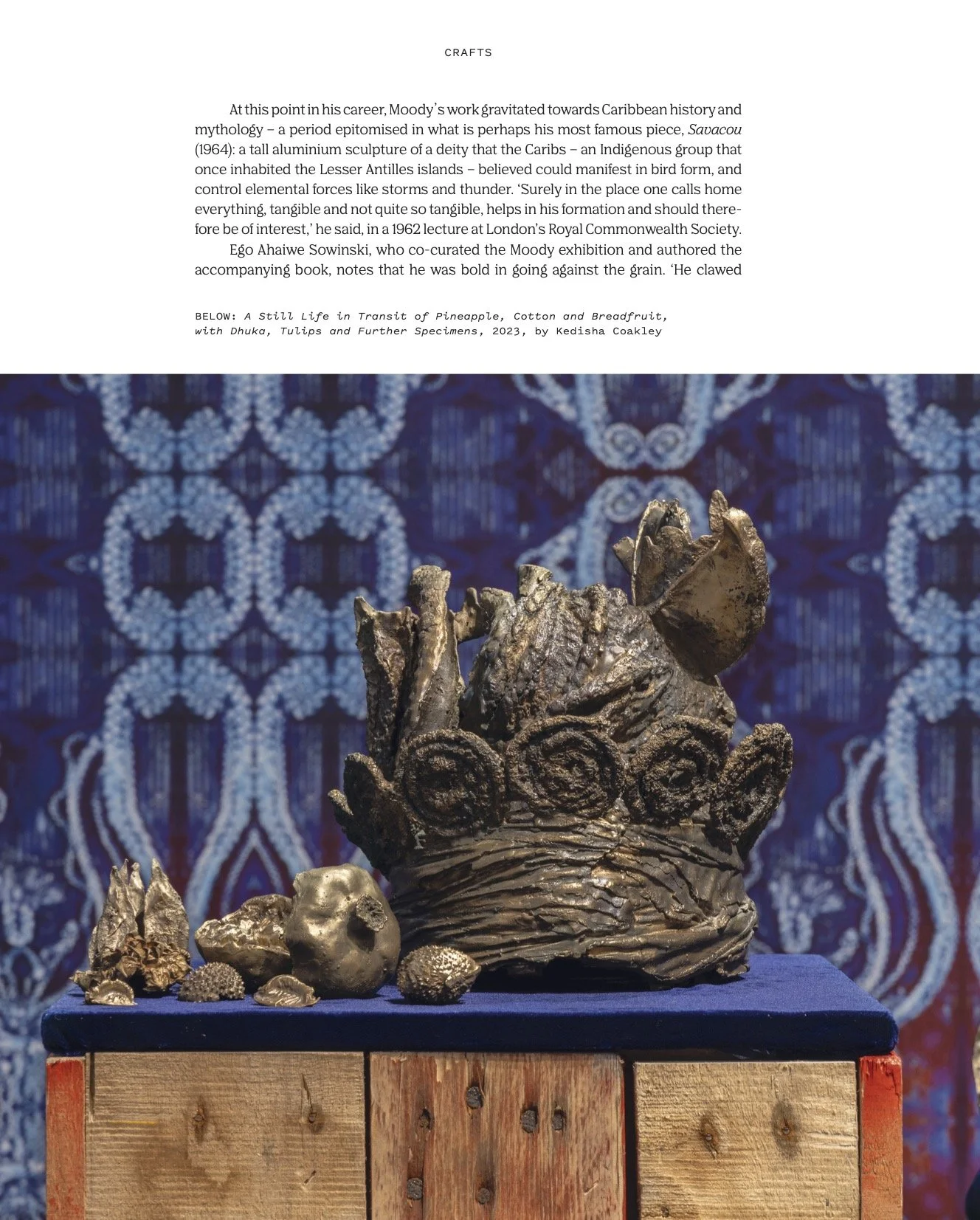 Bronze sculpture of a crab with detailed claws and body, displayed on a wooden pedestal with a blue cloth in front of a patterned blue background.