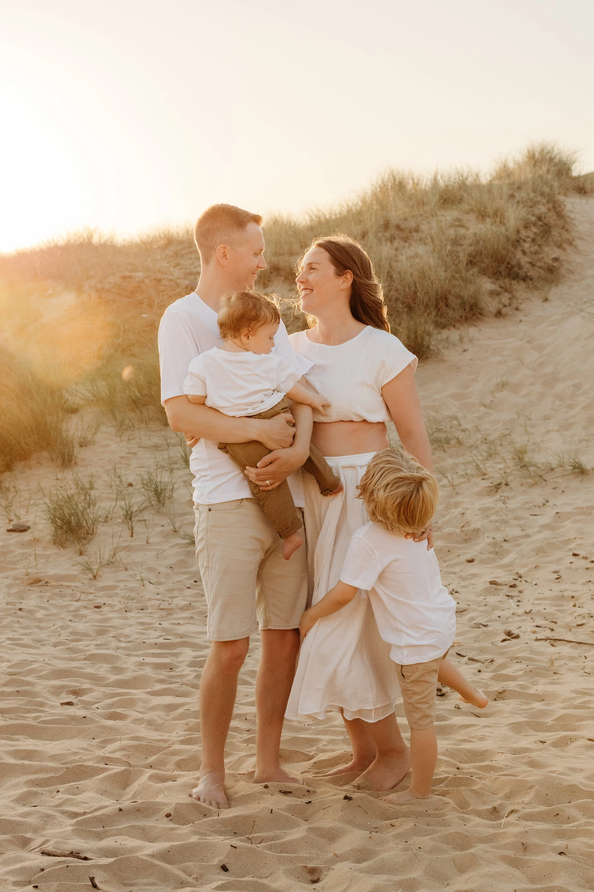 Family on the beach at sunset. Dad holding baby and sun cuddling mum. Parents looking at each other lovingly..