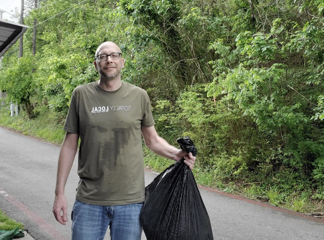 A man with glasses and a beard standing on a rural road, holding a large black trash bag, with greenery and trees in the background.
