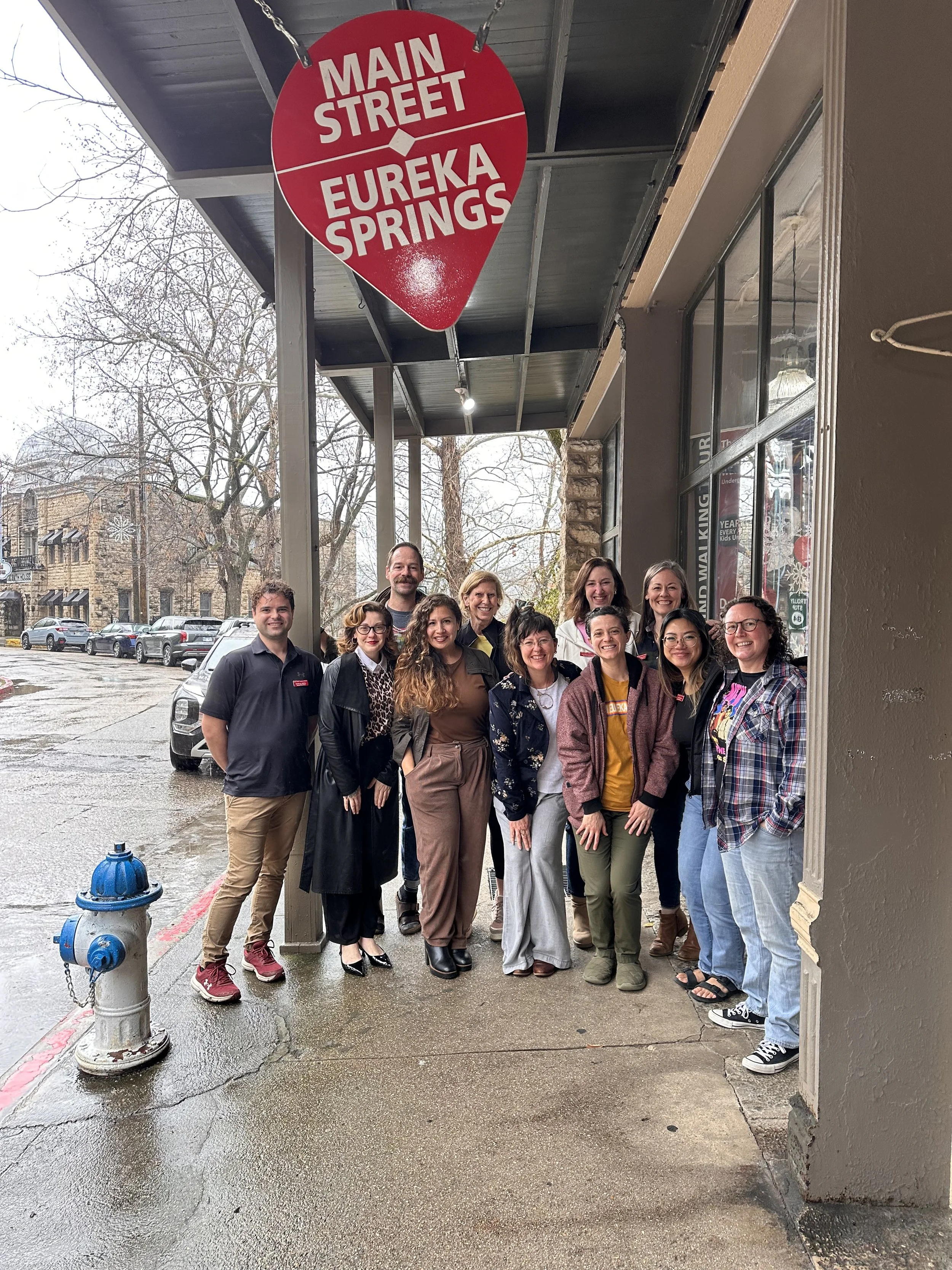 Group of people standing outside a building under a red sign that reads "Main Street Eureka Springs" on a rainy day.