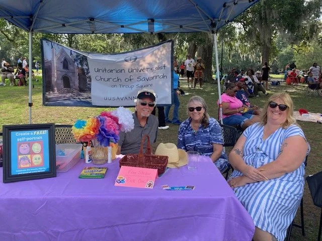 Three women sit at a table under a tent at an outdoor event, with a banner for the Unitarian Universalist Church of Savannah in the background, and other people in the park.
