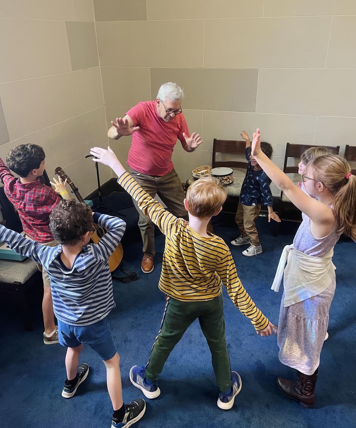 A man with gray hair and glasses is dancing or leading an activity in a room with six children, some of whom have their hands raised. The children are wearing casual clothes, and some are holding musical instruments like guitars and tambourines. The setting appears to be a music or activity room with blue carpet and beige walls.