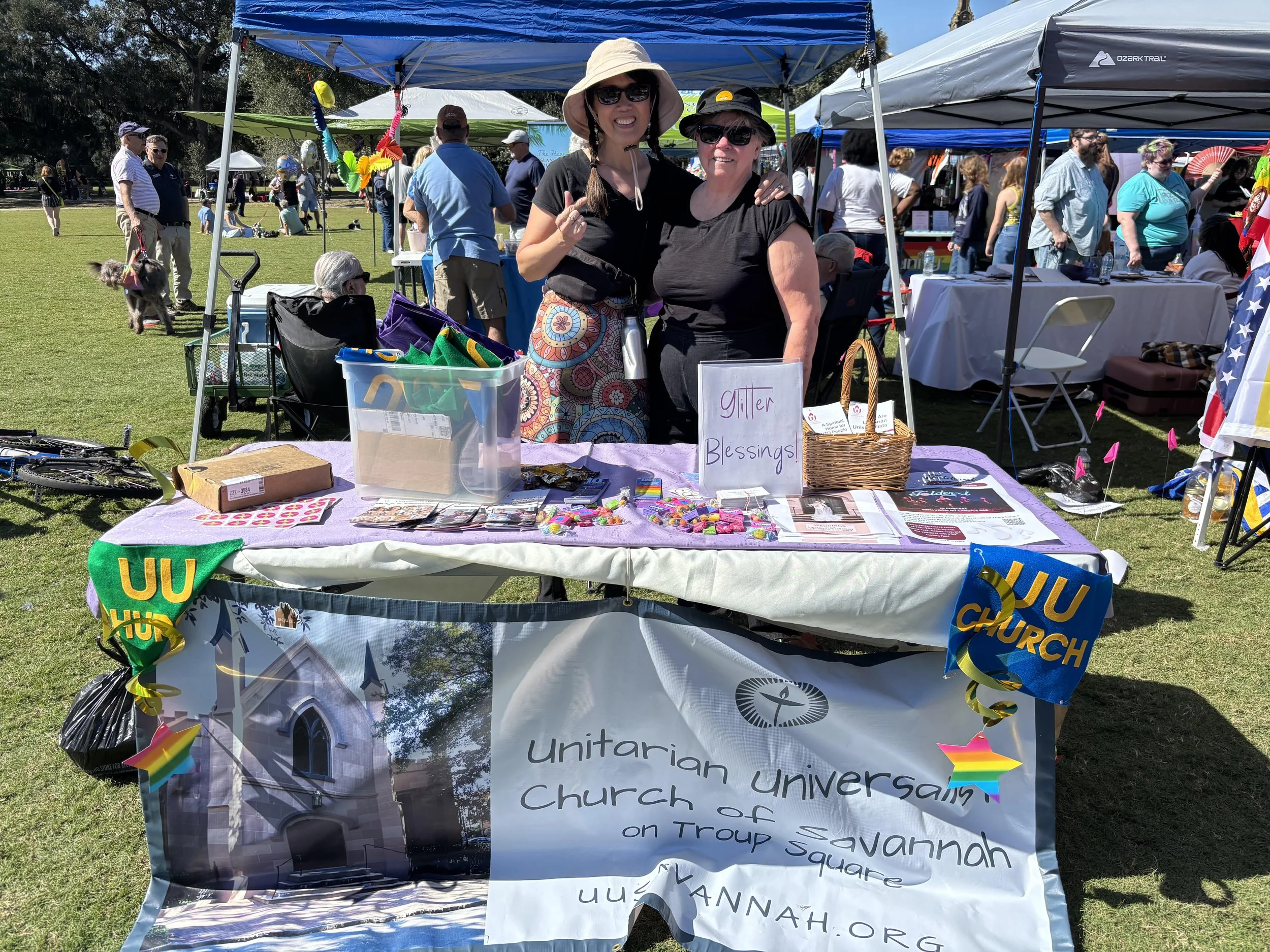Two women standing behind a table at an outdoor event, promoting the Unitarian Universalist Church of Savannah. The table displays colorful items and a sign that says 'Glitter Blessings.' Various tents and people are in the background on a sunny day.
