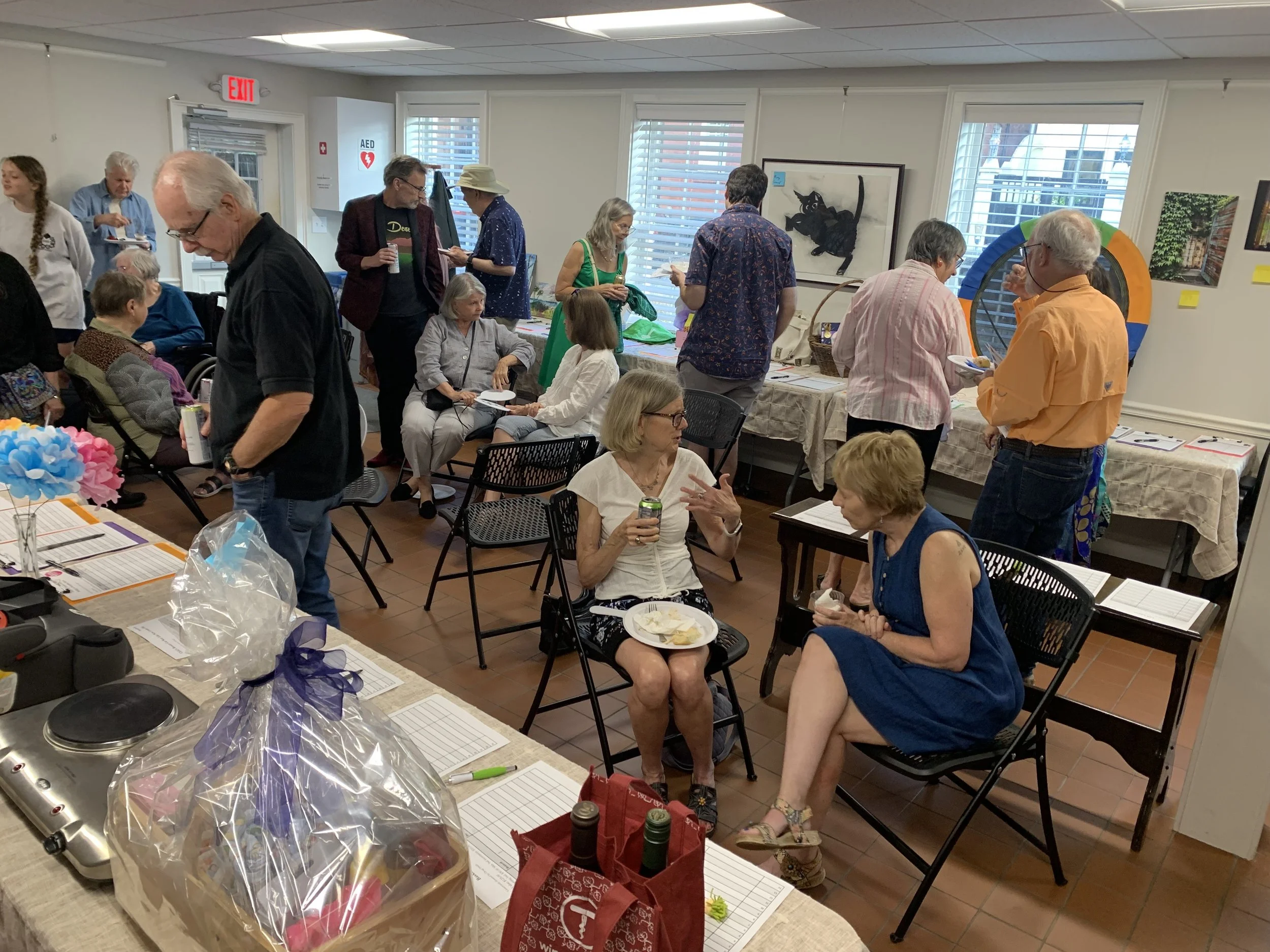 People socializing and talking in an art gallery or community center with tables, artwork on the walls, and a variety of casual and semi-formal attire.