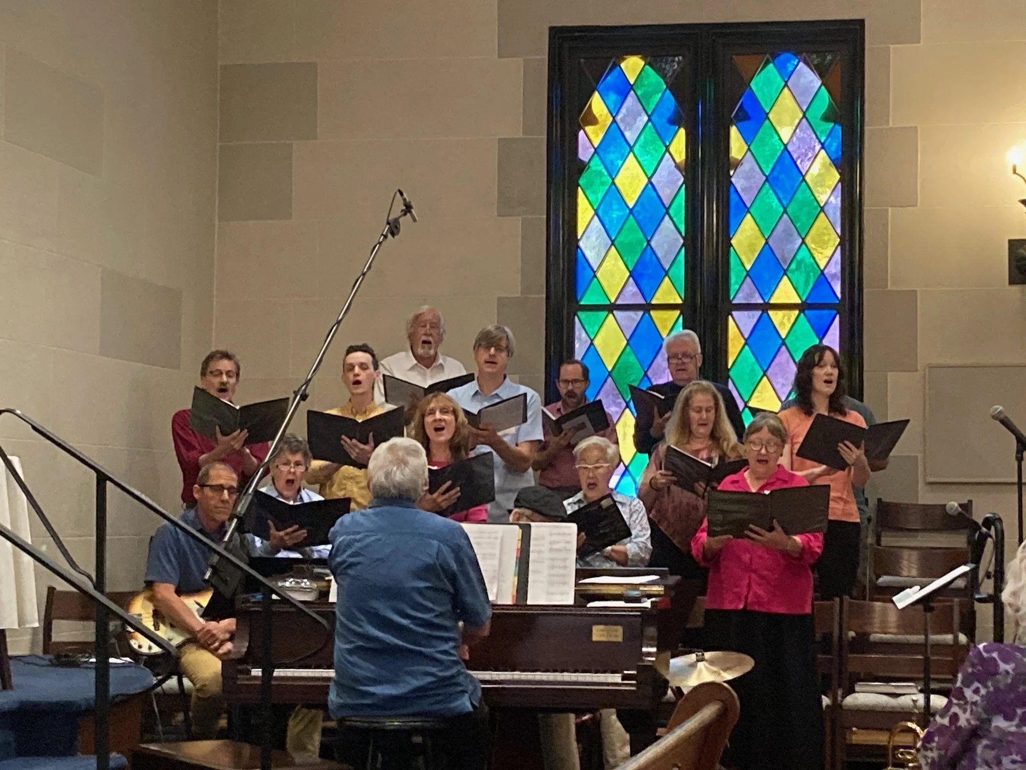 A choir group performing inside a church, with a man playing the piano and a woman conducting, in front of a large stained glass window.