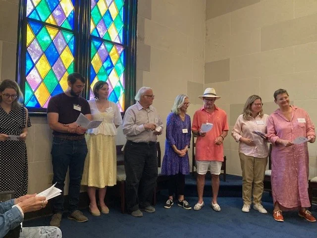Group of seven people standing in a row inside a church or chapel with colorful stained glass windows behind them, holding papers and participating in a group activity or ceremony.