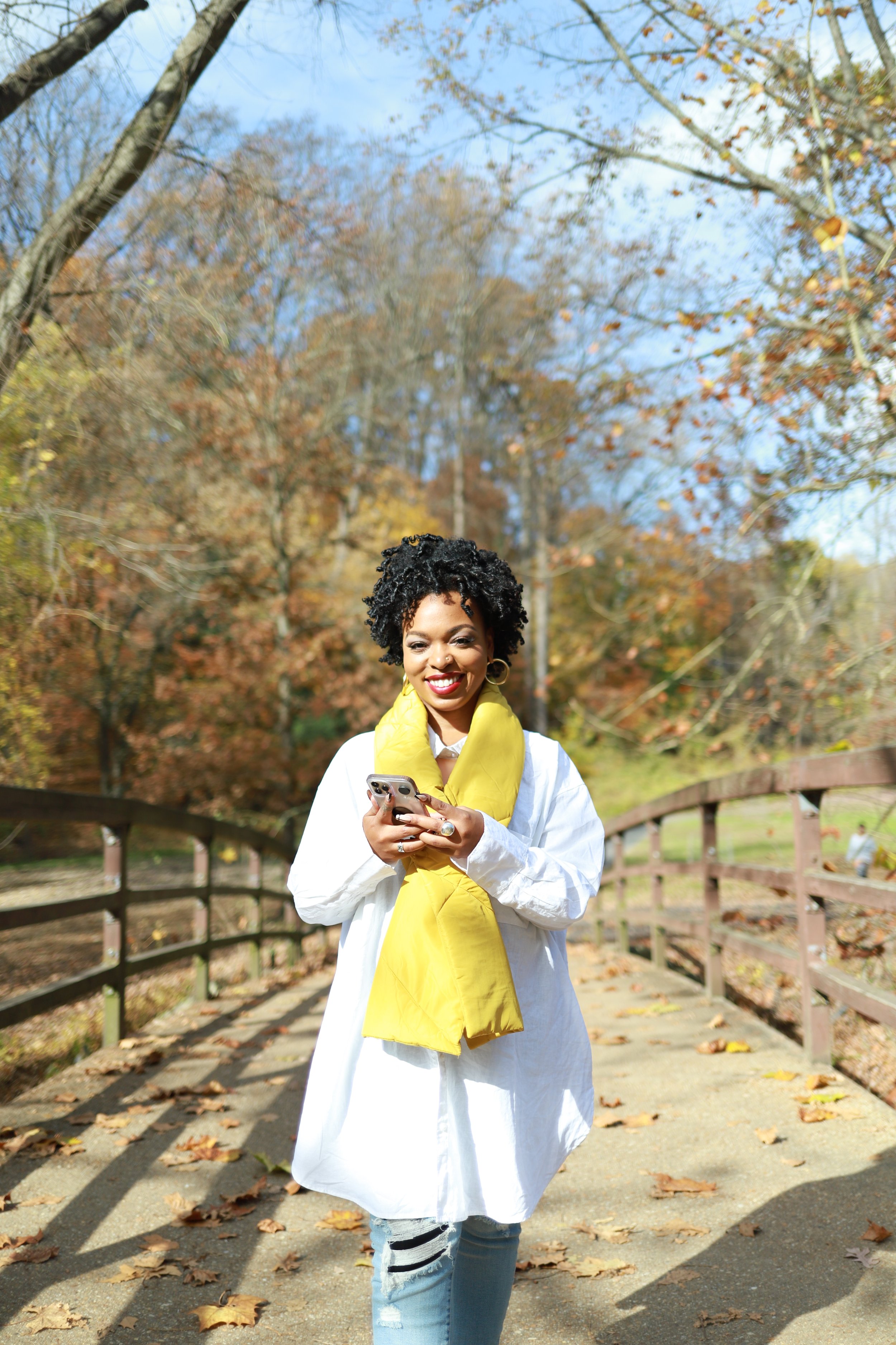 Ebony stands on a bridge wearing a white blouse and yellow scarf. She is smiling and holding her cell phone on an autumn day with leaves and sunshine.