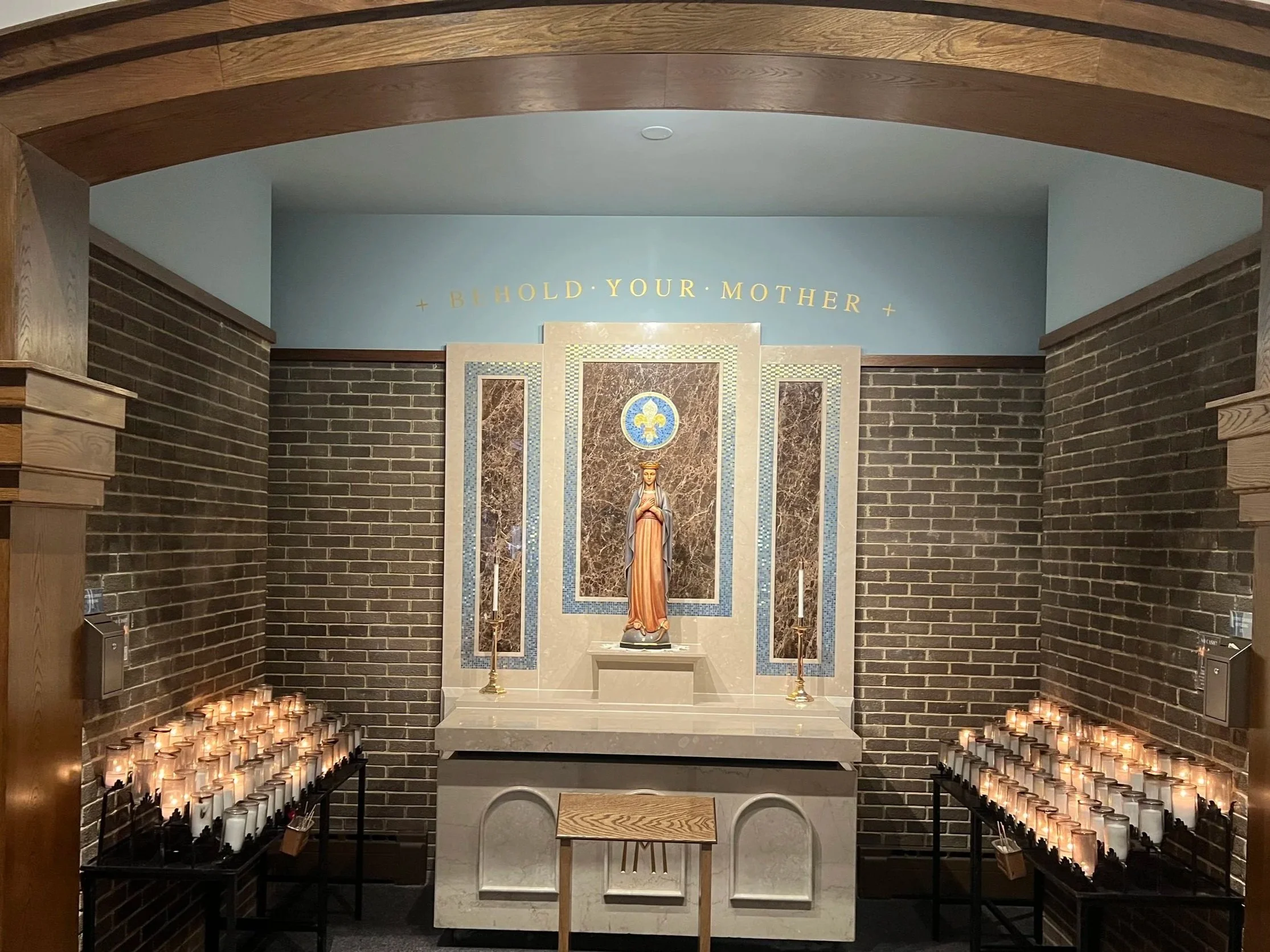 A religious altar with a statue of the Virgin Mary, candles on both sides, and the inscription 'BOLD YOUR MOTHER' on the wall above.