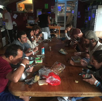 Groupe de personnes assises autour d'une grande table en train de manger et d'utiliser des appareils électroniques dans un endroit convivial et éclairé.