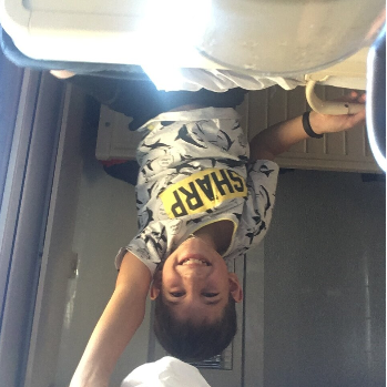 Un garçon souriant dans une cuisine, debout devant un évier, portant un t-shirt camouflage avec la phrase 'SHARP'