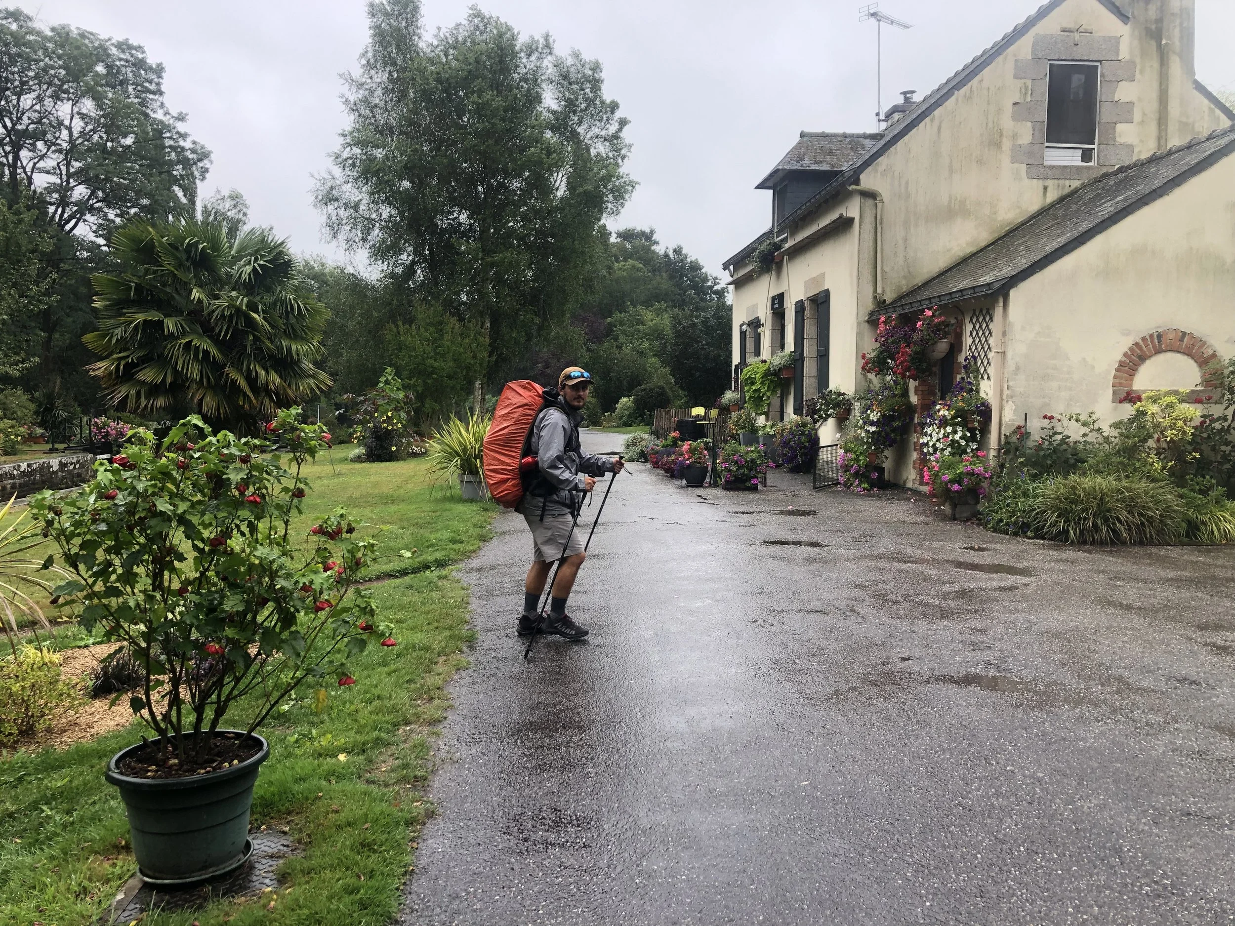 Un randonneur avec un grand sac à dos et des bâtons sur un chemin devant une maison située dans un jardin fleuri par temps pluvieux.