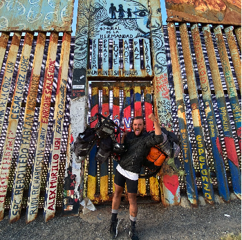 Deux personnes posant devant un mur de mosaïque coloré avec des inscriptions et des motifs, un homme tenant un sac de voyage et un autre portant un sac à dos orange.