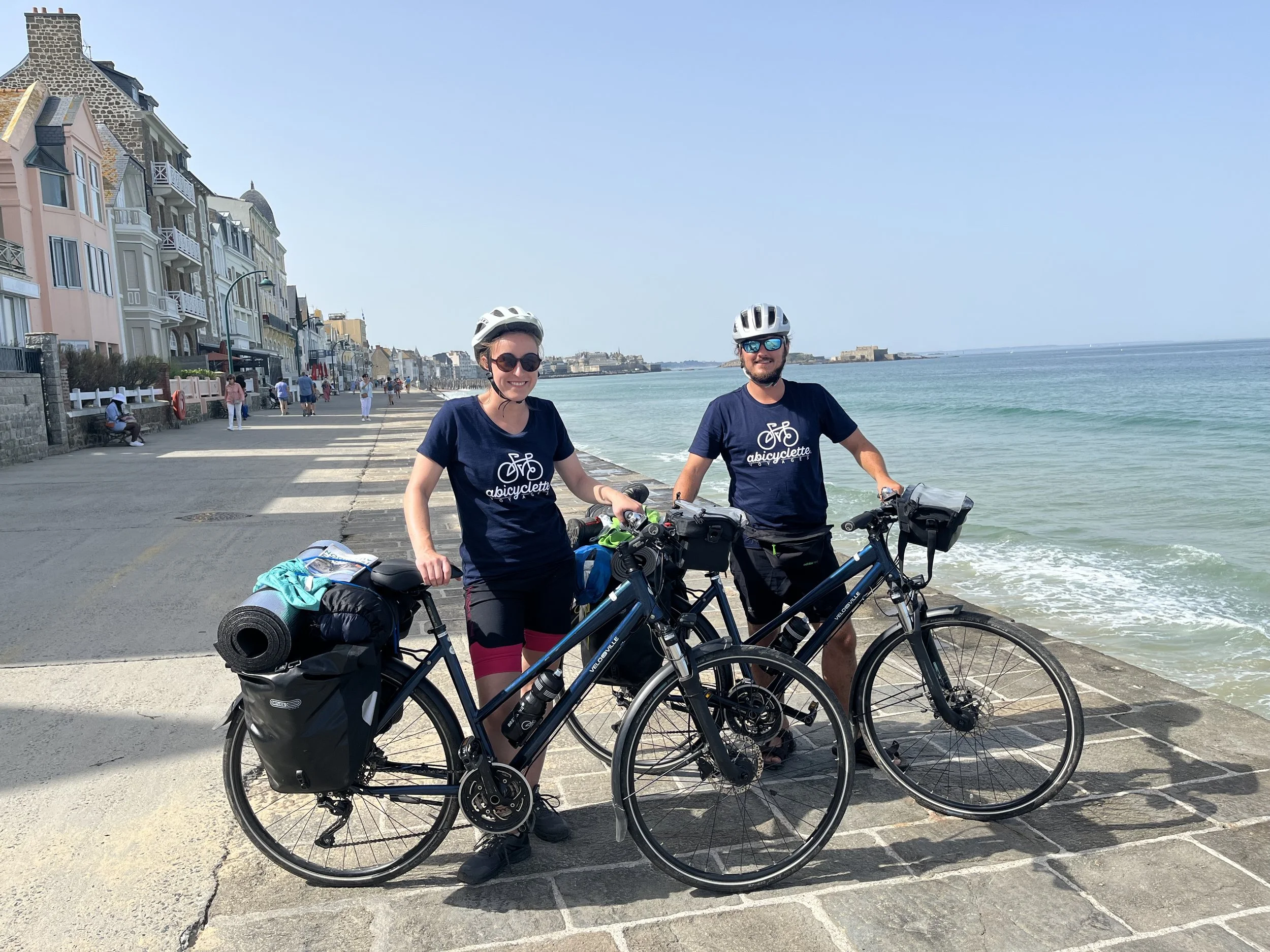 Deux cyclistes portant des casques debout à côté de leurs vélos sur une promenade au bord de la mer, avec des bâtiments résidentiels en arrière-plan et une mer calme sous un ciel bleu.