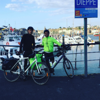 Deux personnes en vêtements de cyclisme posent avec leurs vélos devant la marina de Dieppe, avec des bateaux et des bâtiments en arrière-plan.